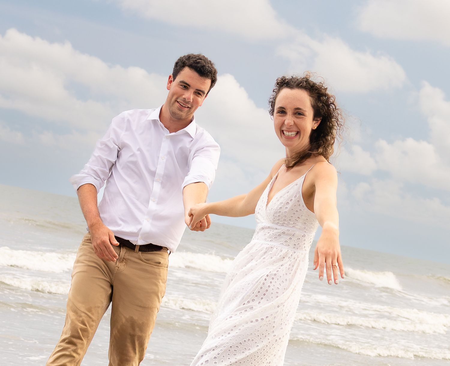 Happy couple holding hands and walking on a sunny beach with ocean waves, woman in white dress, man in white shirt.