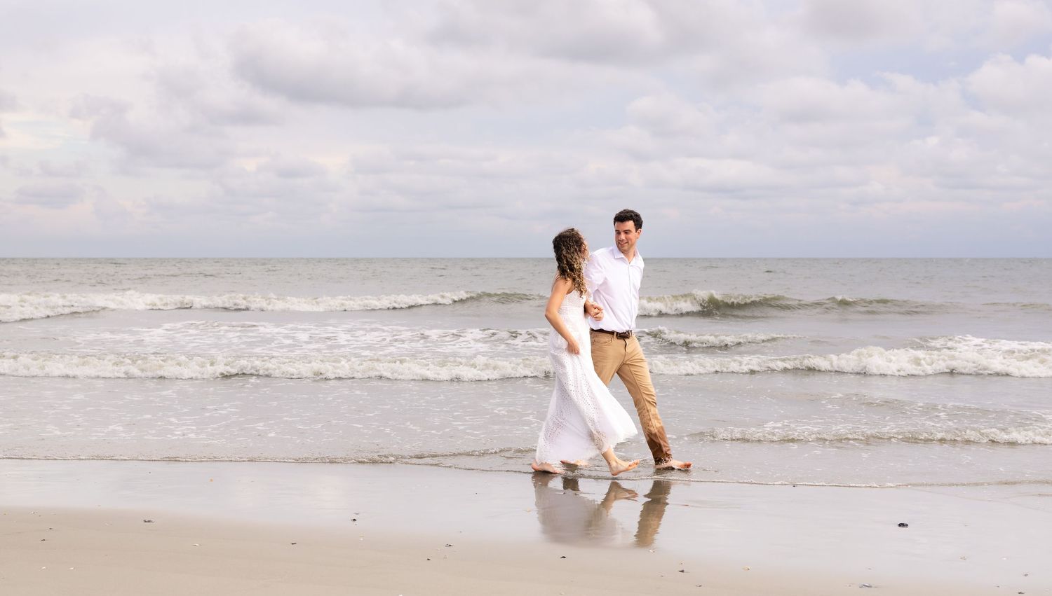 Couple in white and beige outfits walking hand-in-hand on a sandy beach with ocean waves in the background.