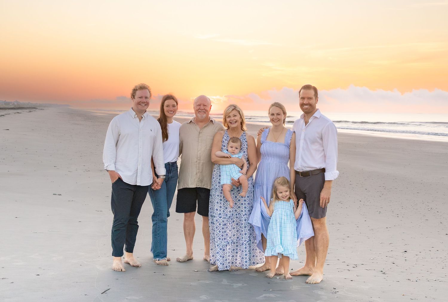 Family group photo on beach at sunset, smiling adults and children posing together by ocean waves.