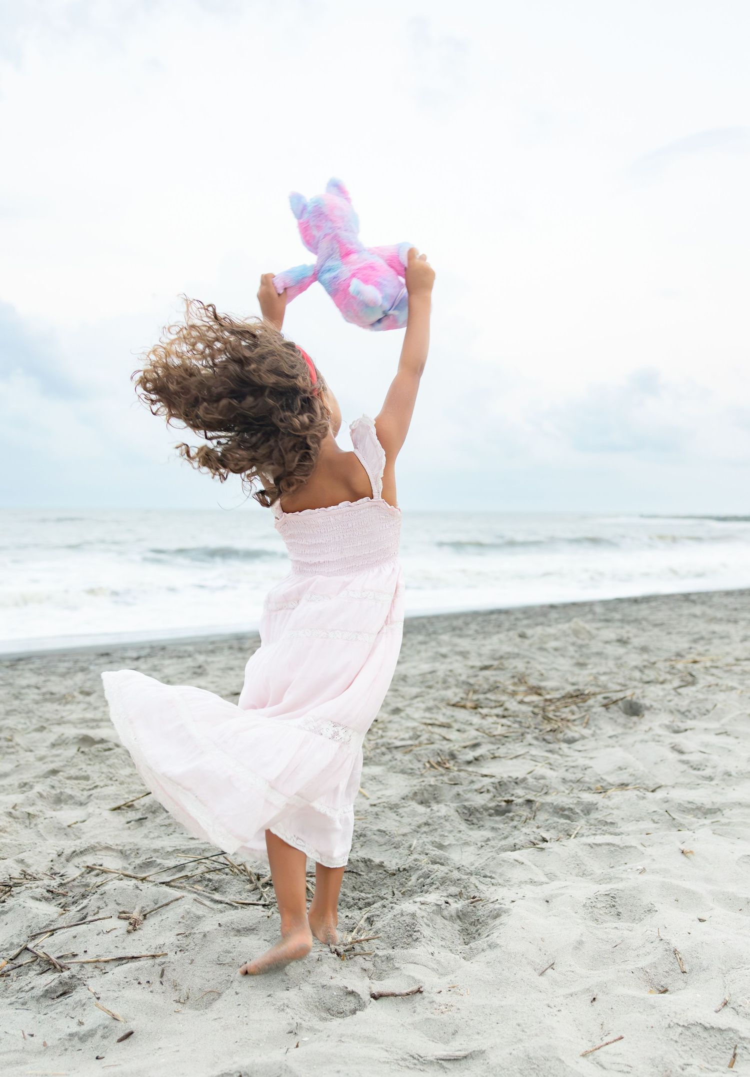 Young girl in pink dress dancing on beach, holding colorful stuffed animal aloft with curly hair flowing in wind.