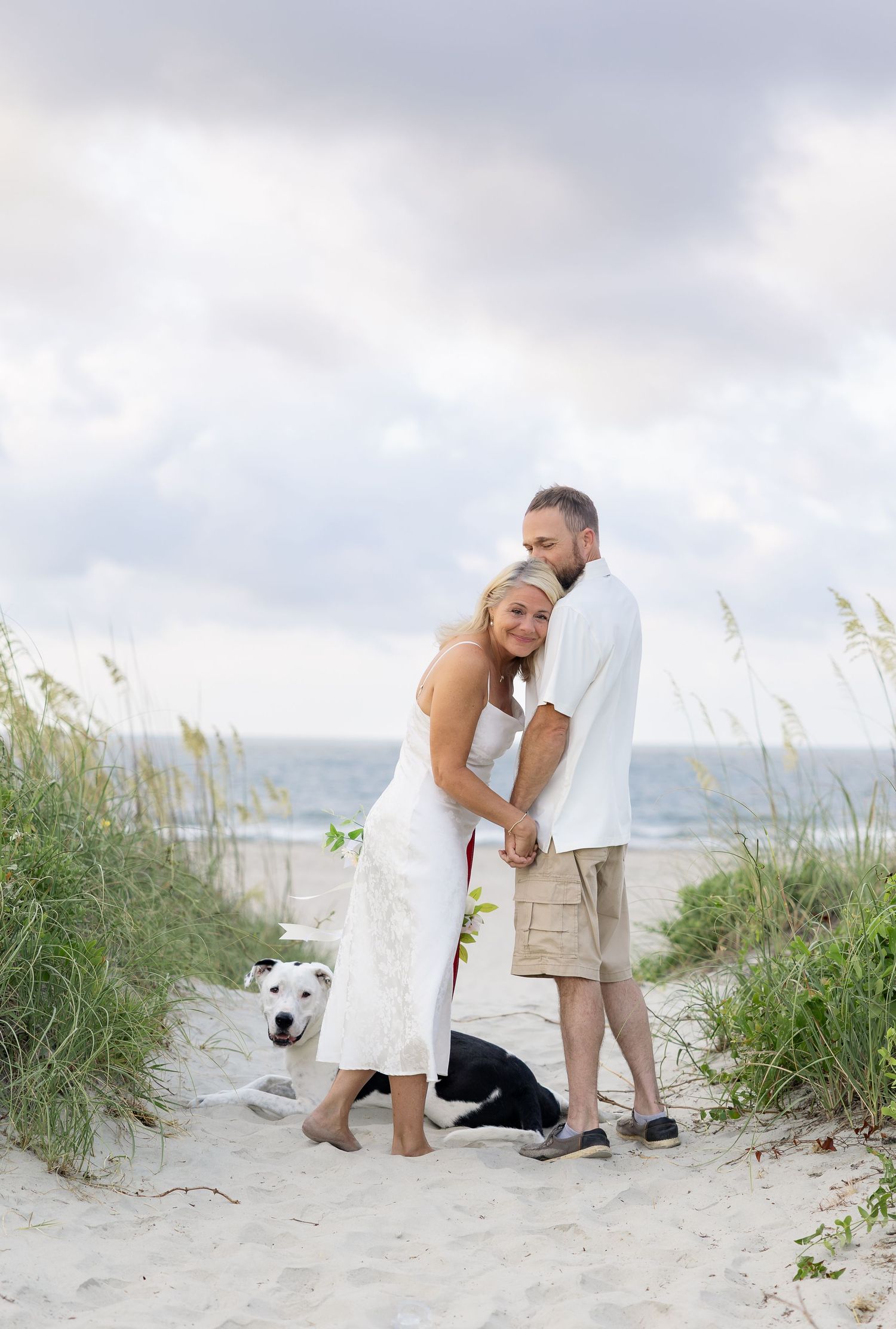 Couple in white wedding attire embracing on sandy beach path with two dogs and sea oats under cloudy sky.
