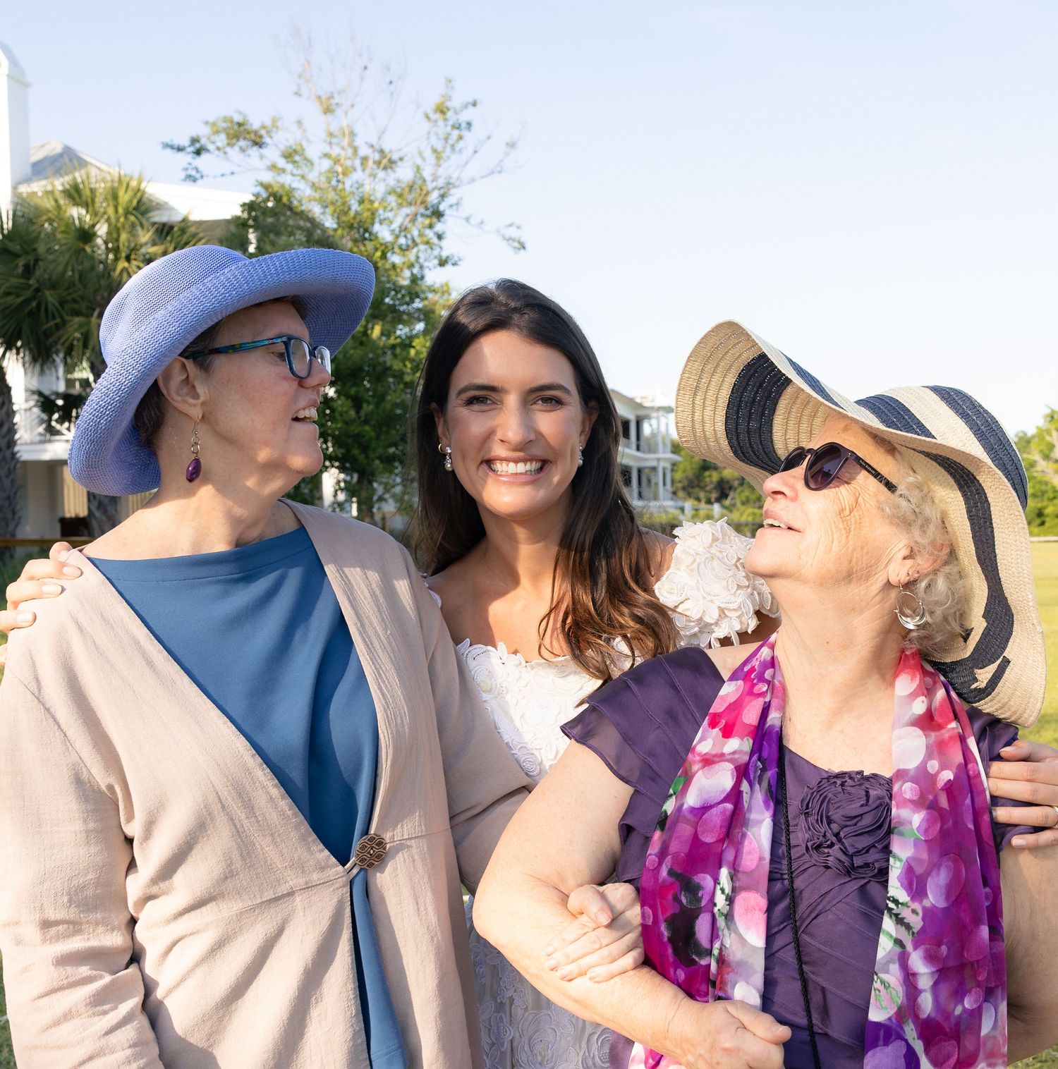 Three women smiling together outdoors at a garden event, two wearing decorative hats.