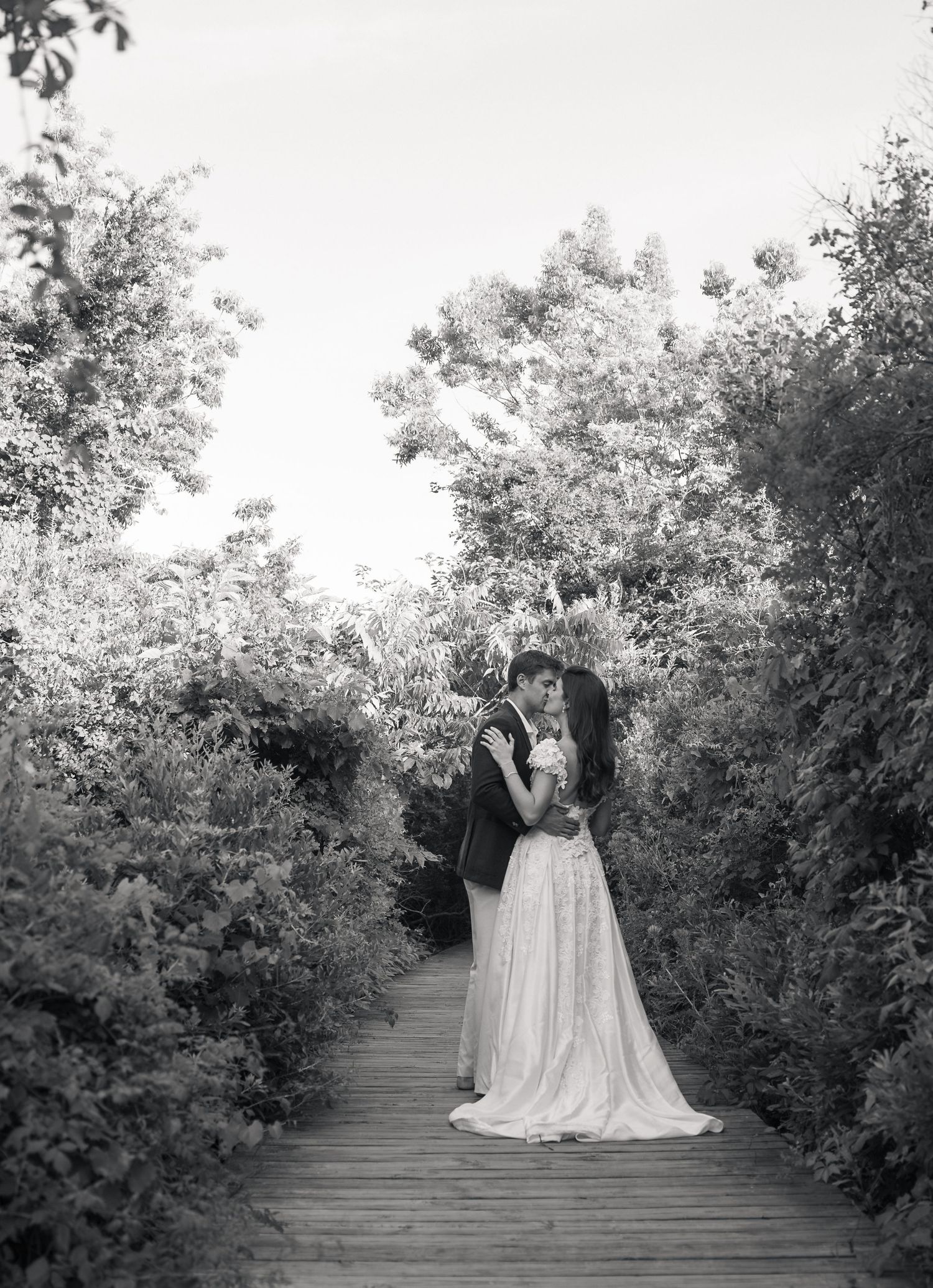 Black and white photo of a couple kissing on a wooden path lined with trees.