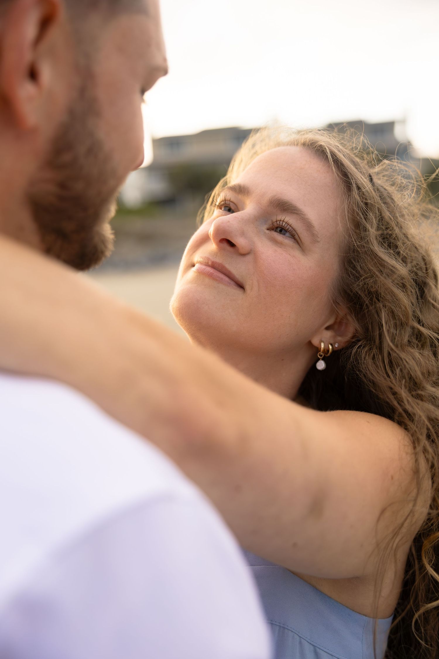 Woman with curly hair gazes lovingly at bearded man during romantic beach engagement photo session.