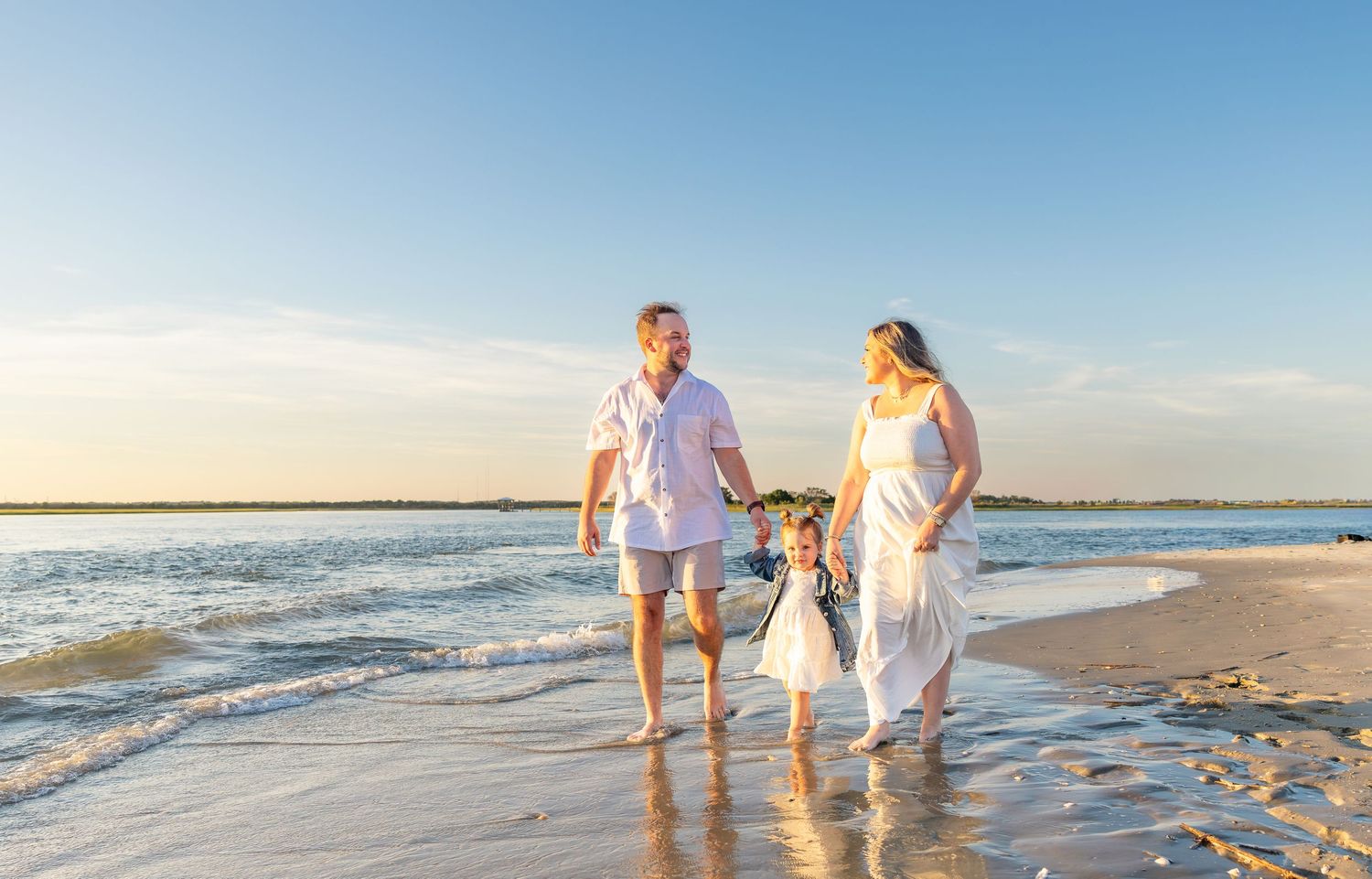 Family of three in white outfits walking on a sunny beach, waves gently lapping at their feet.