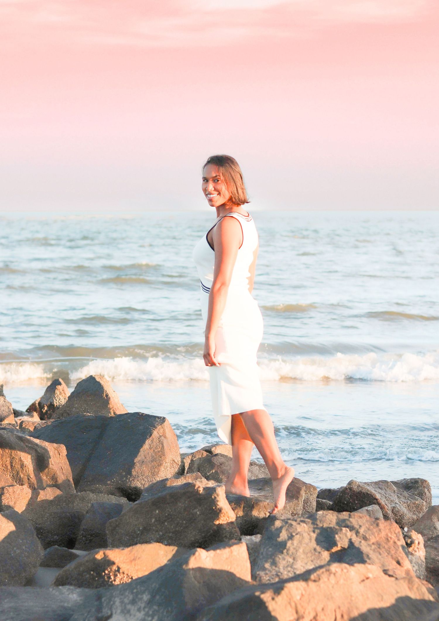Person in white dress walking on rocky coastal shoreline during sunset.
