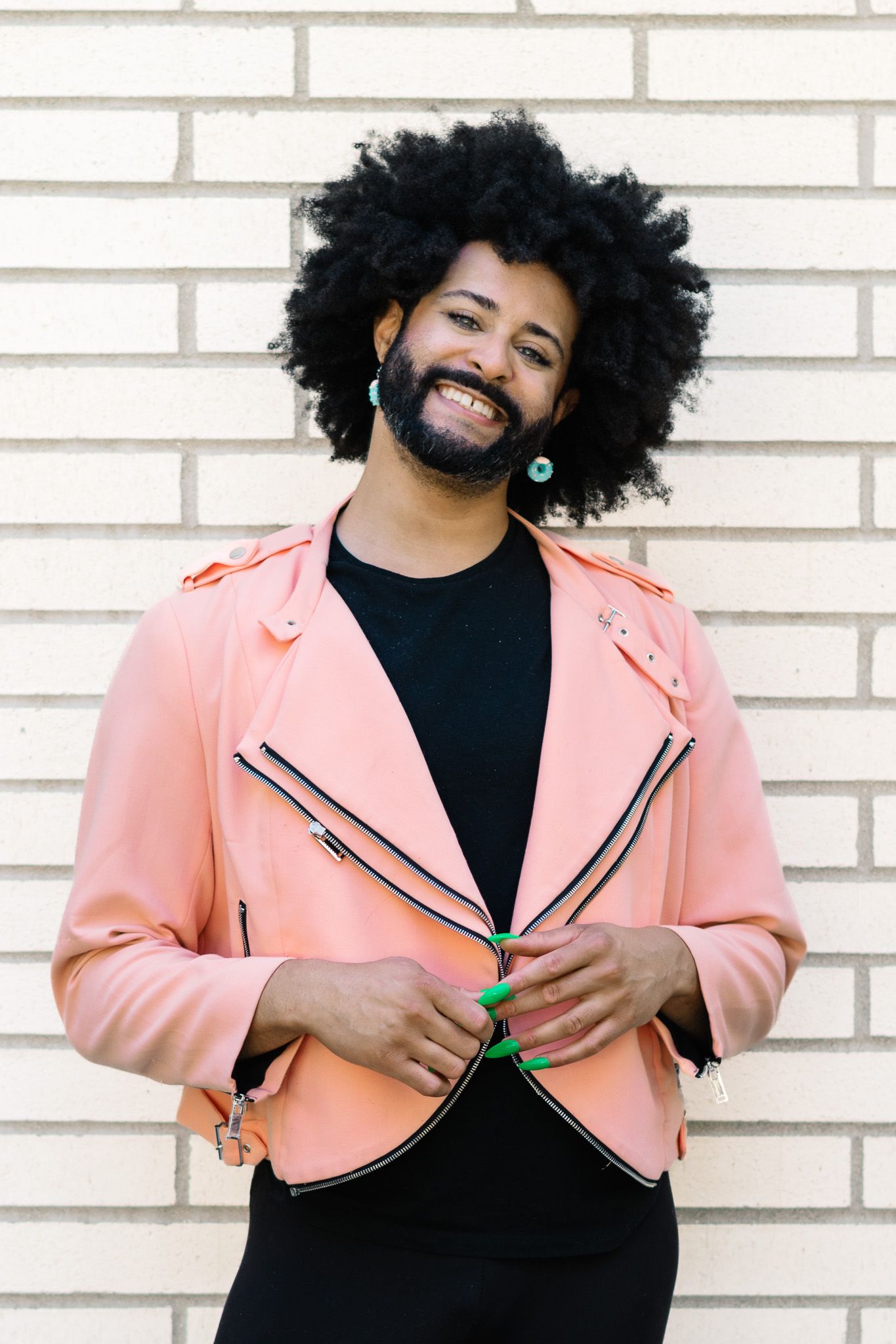 Cheerful person with natural afro hair wearing a pink leather jacket against a white brick wall.
