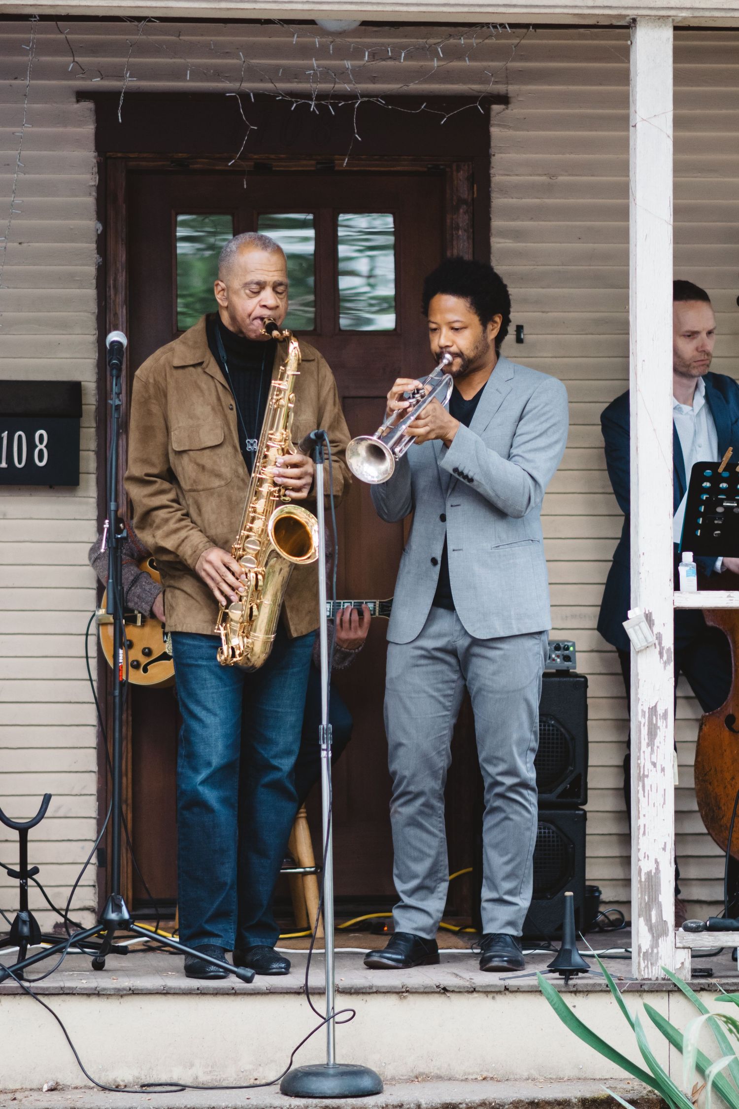 Musicians performing with saxophones and trumpets on a rustic porch of a house numbered 108.