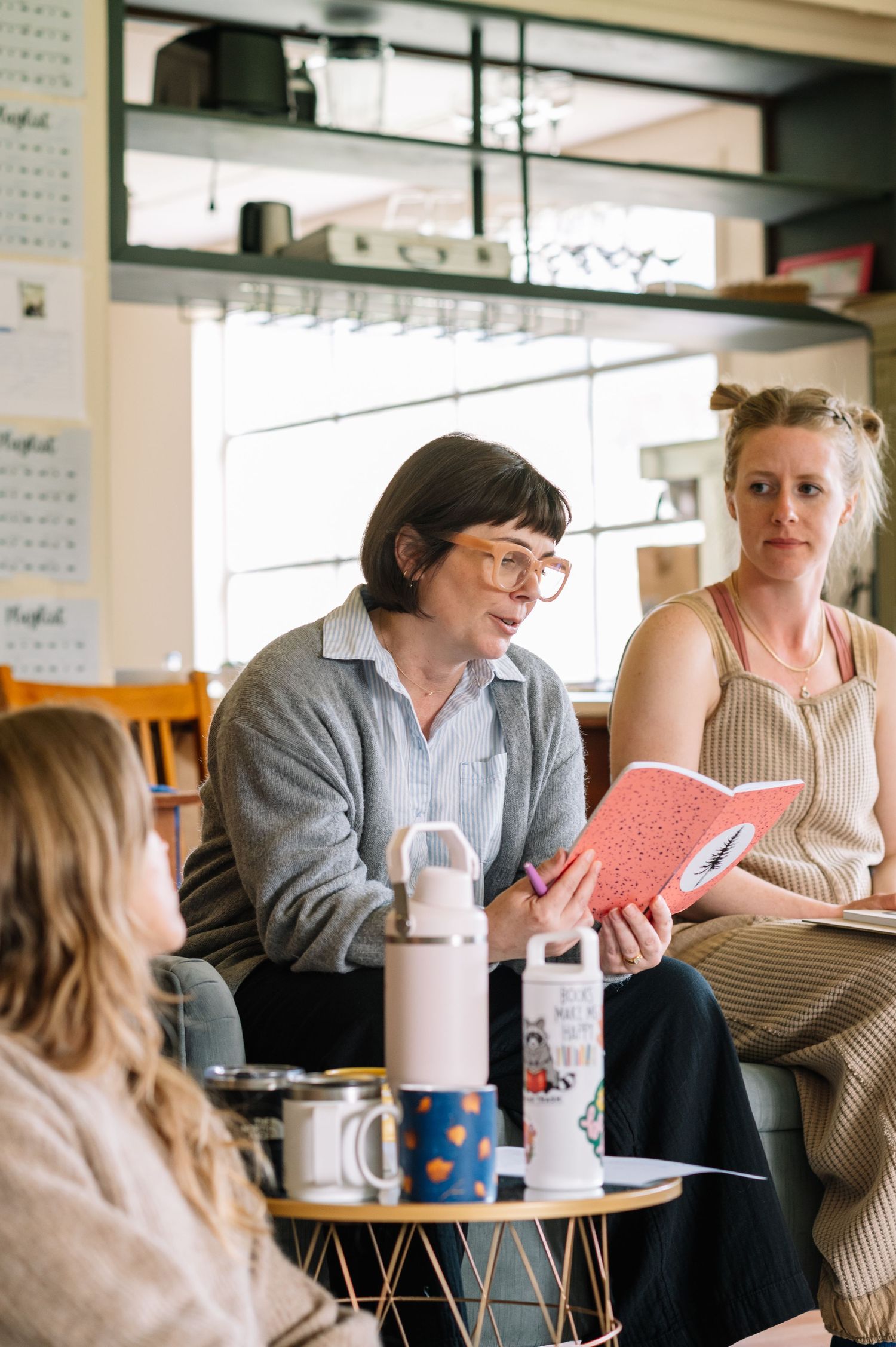 Three women gathered in a cozy cafe, one holding a pink notebook during an engaging book club discussion.