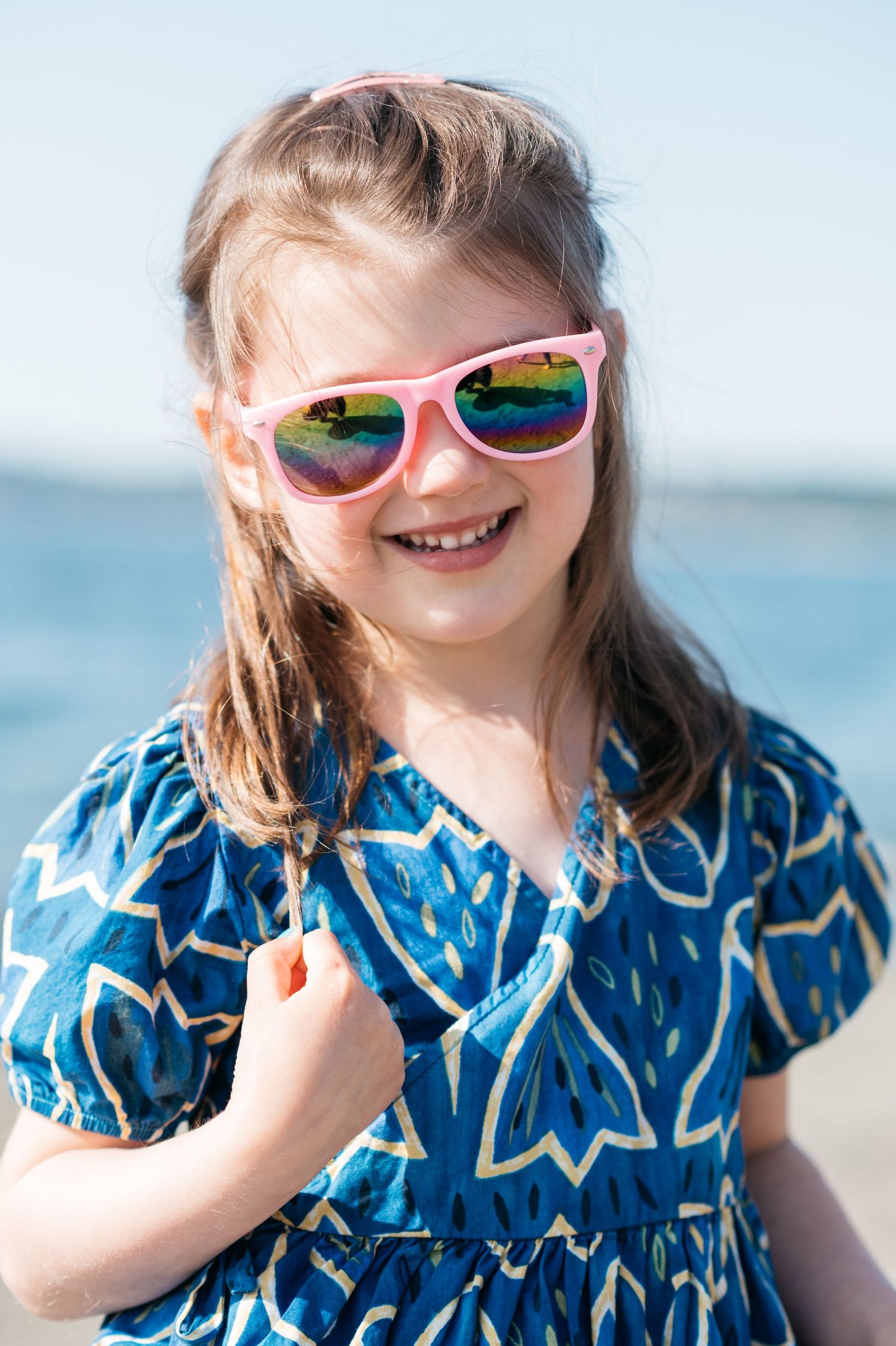 Child wearing pink sunglasses and blue dress smiles at the beach.
