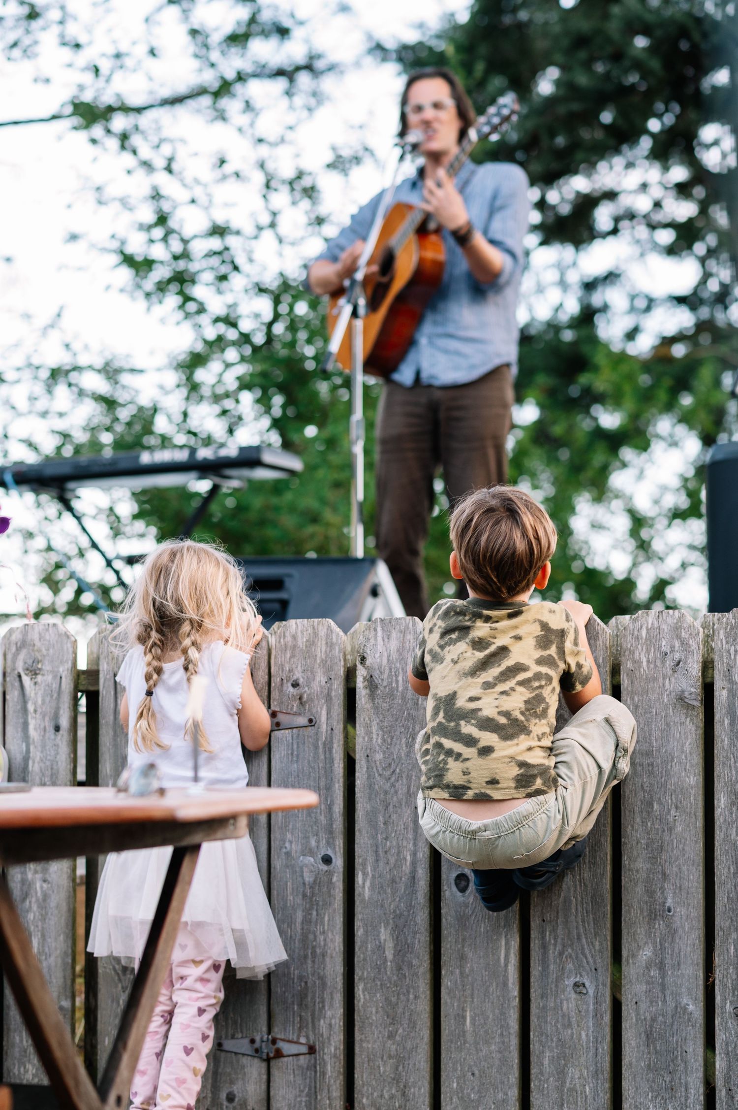 Two children watch a man playing acoustic guitar on an outdoor stage over a wooden fence.