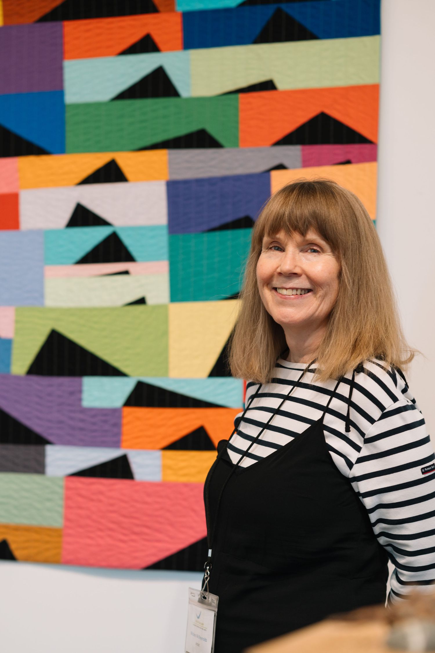 A person in a striped shirt sits in front of a colorful geometric quilt display at an art exhibition.
