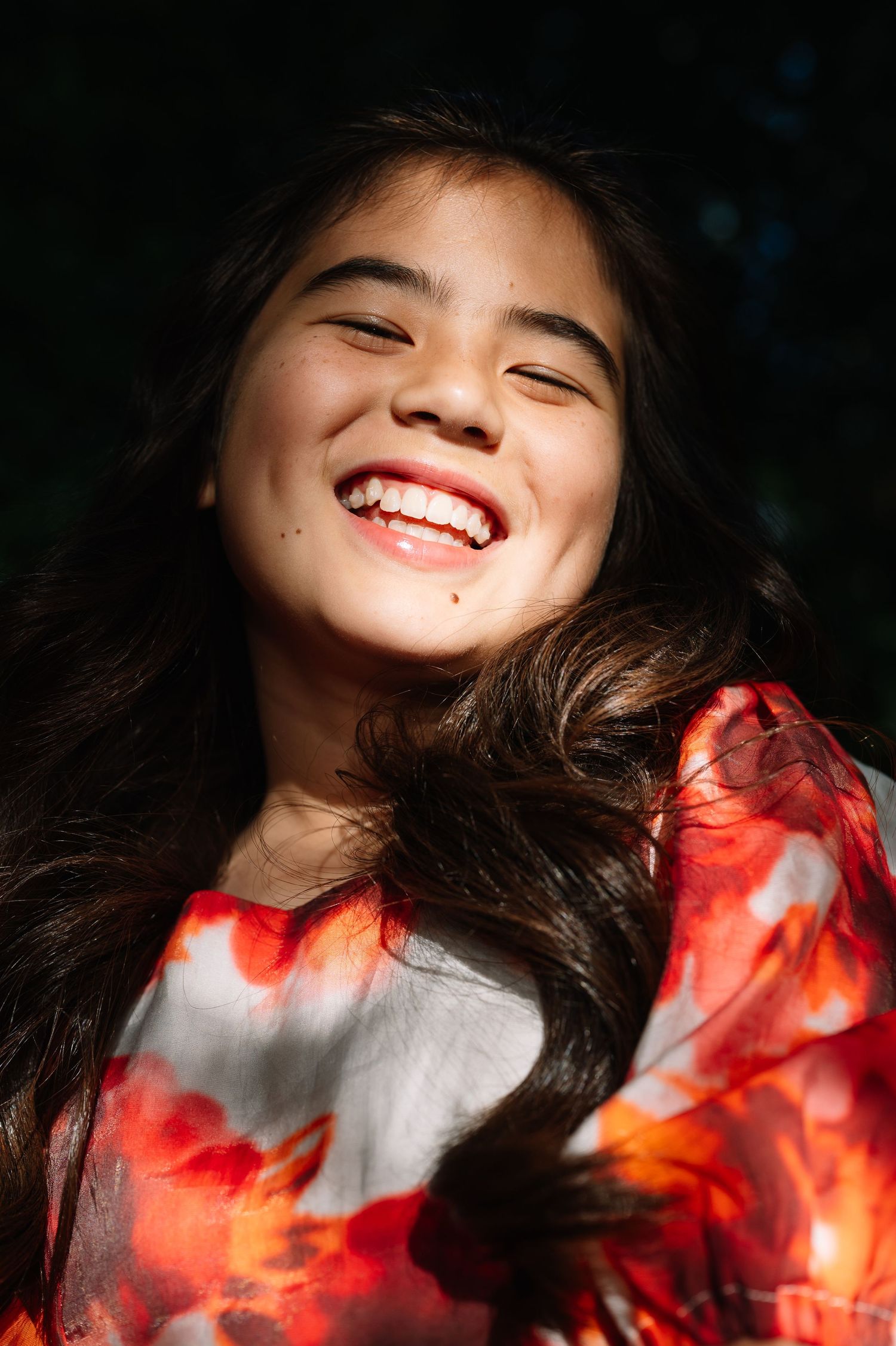 Joyful young woman with long dark hair laughing in sunlight, wearing a red floral dress against dark background.