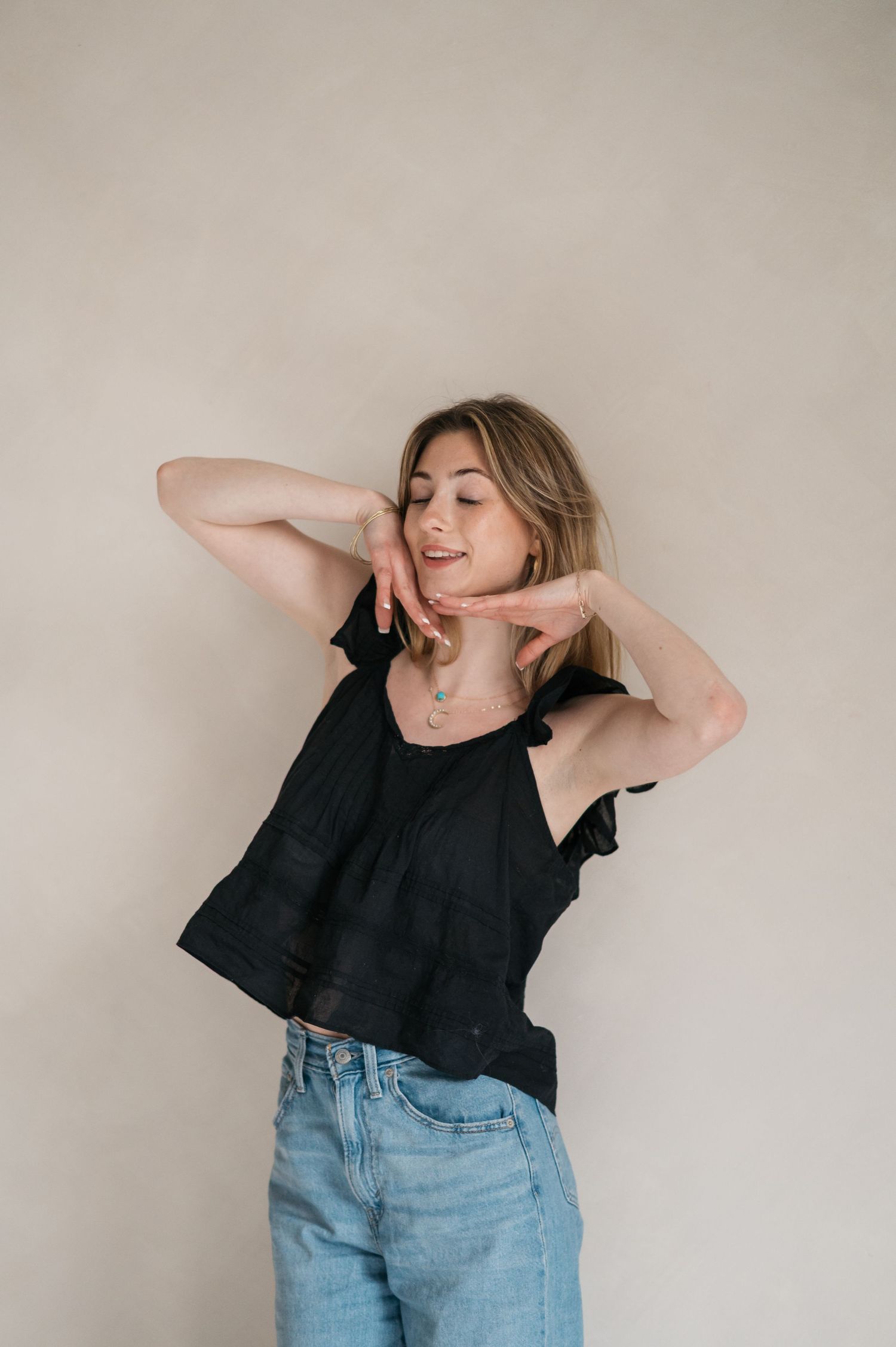 Person in black ruffled top and jeans posing playfully against neutral wall.