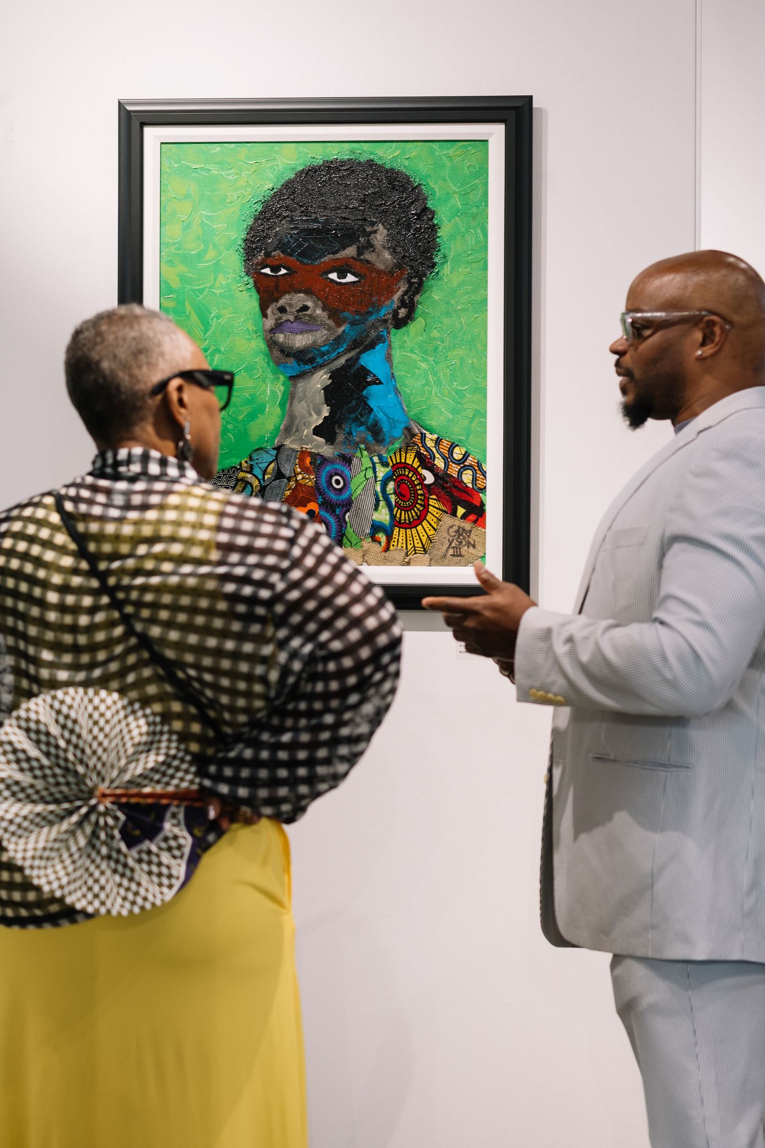 Gallery visitor in checkered shirt and yellow pants examining framed artwork on white wall.