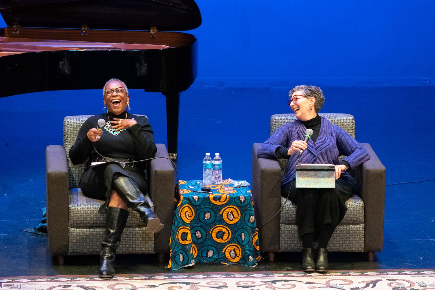 Two people sit in dark chairs on a blue stage having an animated discussion during an event.