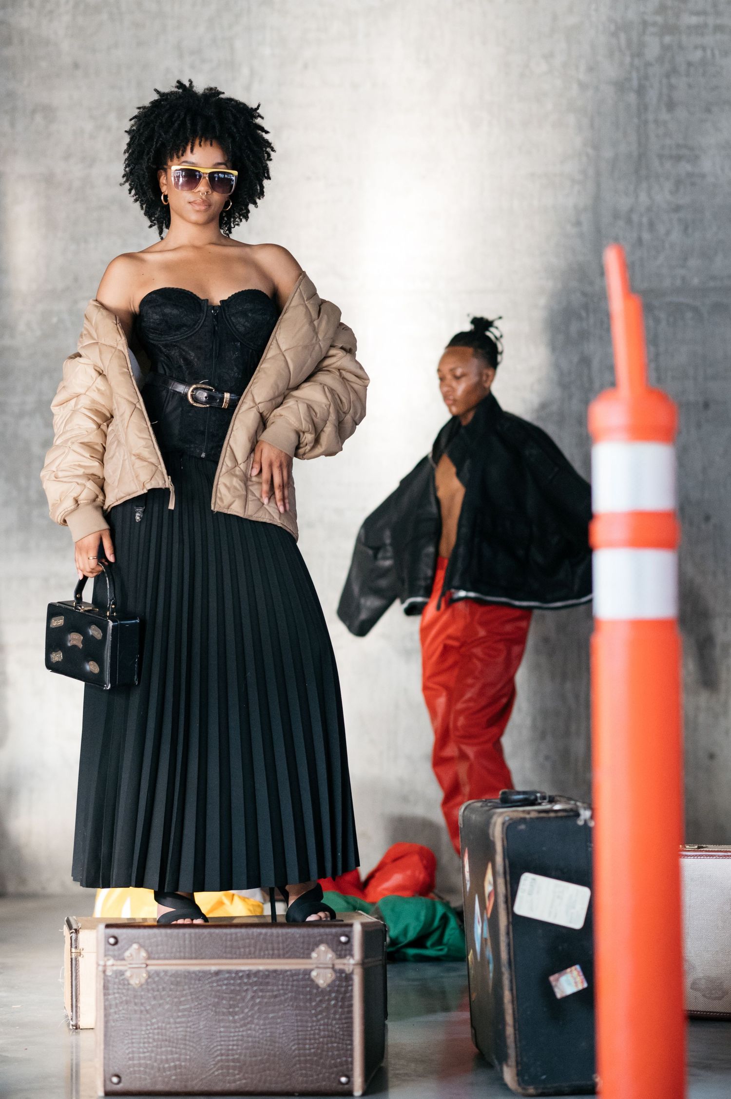 Fashion photoshoot with a black dress, beige jacket, and orange traffic cone against an industrial backdrop.