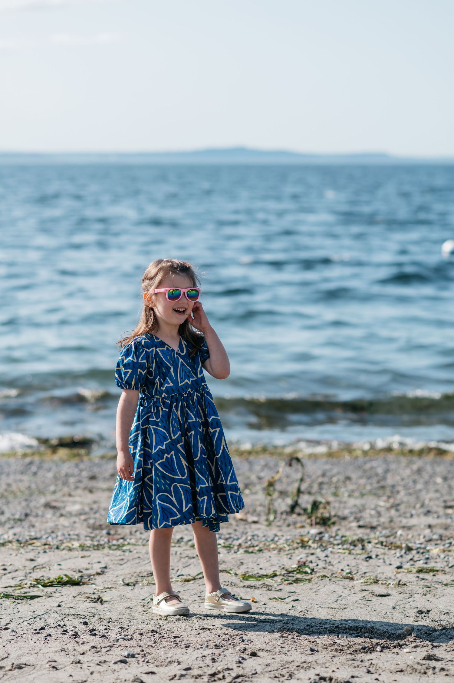 Child in blue patterned dress plays joyfully by the ocean.