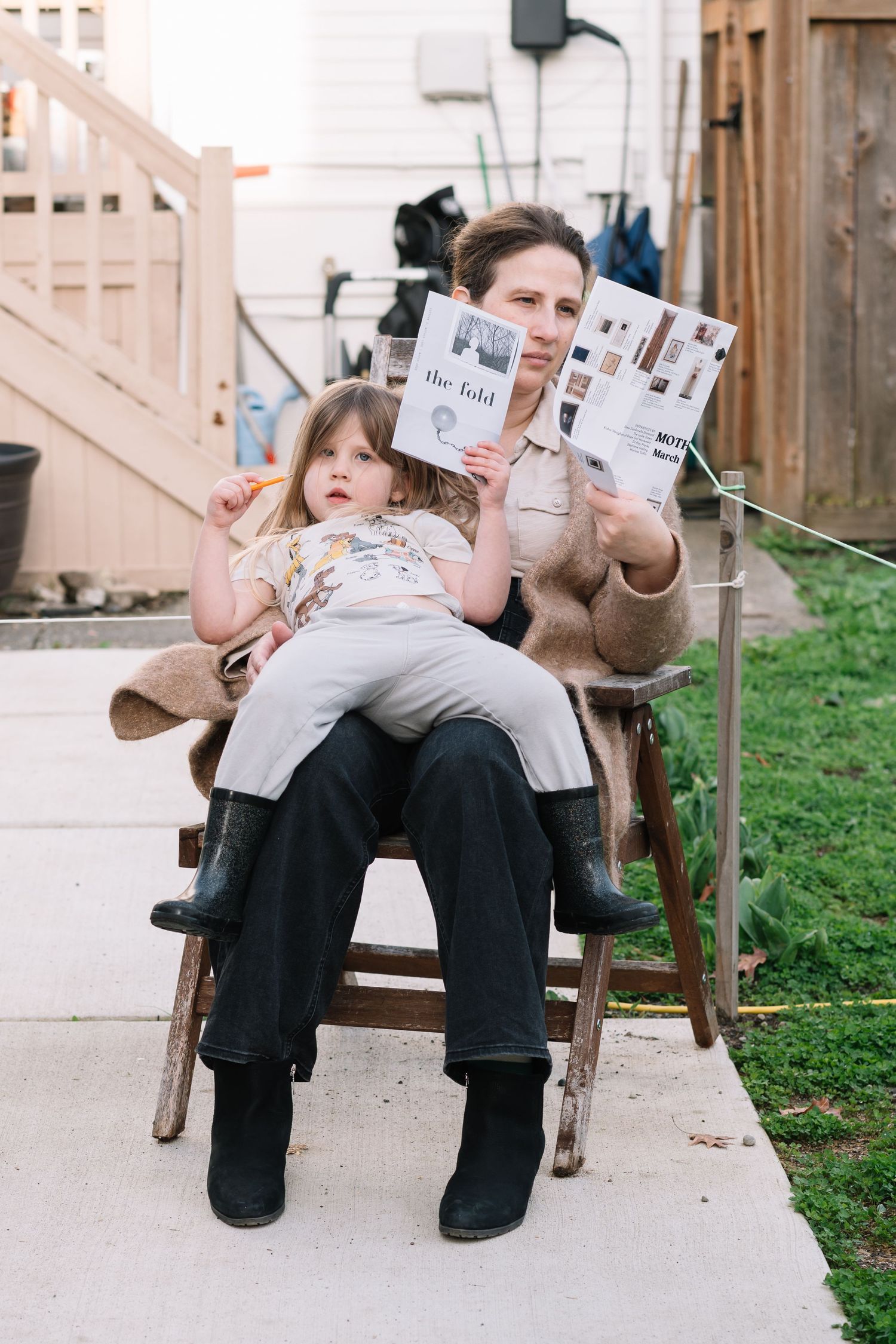 Adult and child sitting in outdoor chair reading a magazine together in a backyard setting.