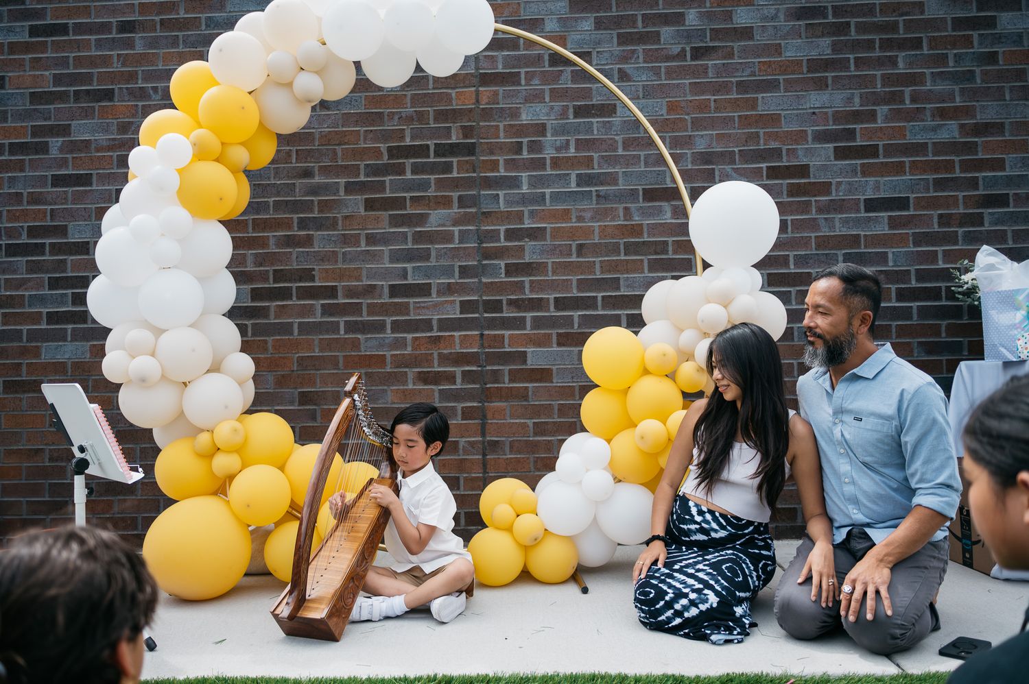 Party setting with white and yellow balloon arch decorations against brick wall.