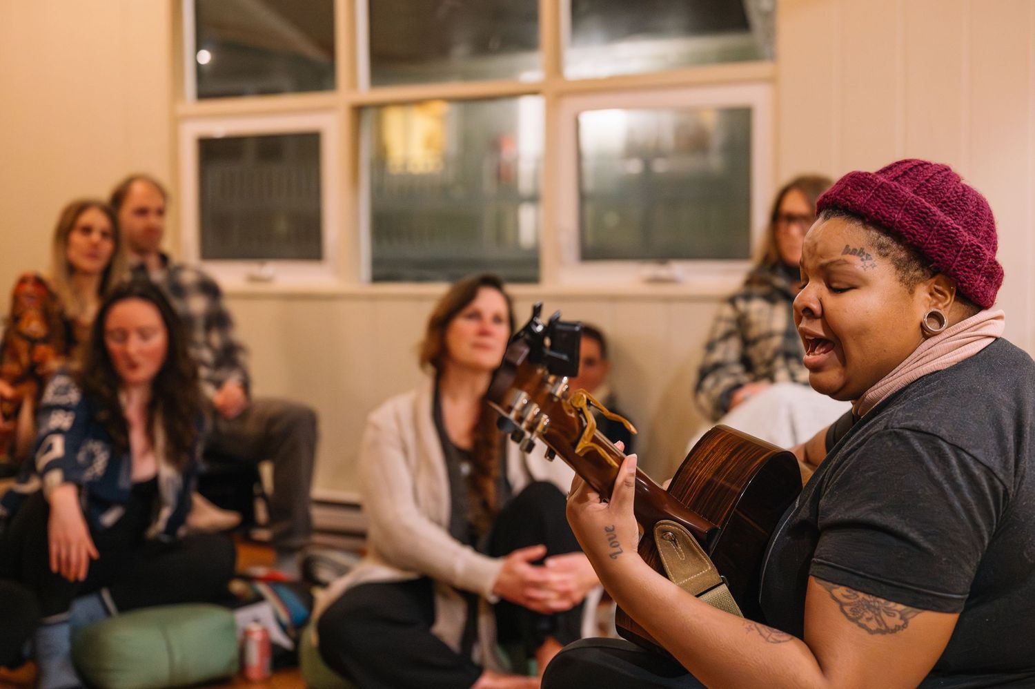 Woman in red beanie plays acoustic guitar while singing for an attentive group seated in a cozy indoor gathering.