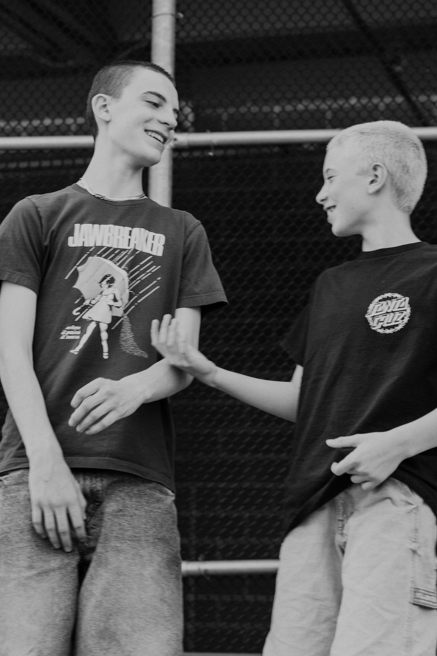 Two smiling teens in graphic tees, one Jawbreaker and one Santa Cruz, interact cheerfully near a chain-link fence.