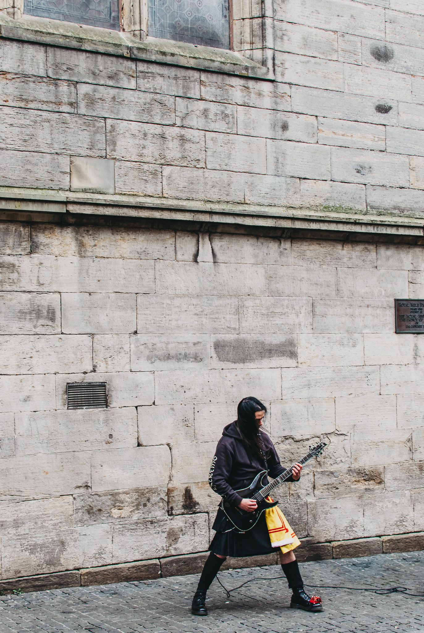Person in dark clothing carrying a guitar walks past weathered stone wall.