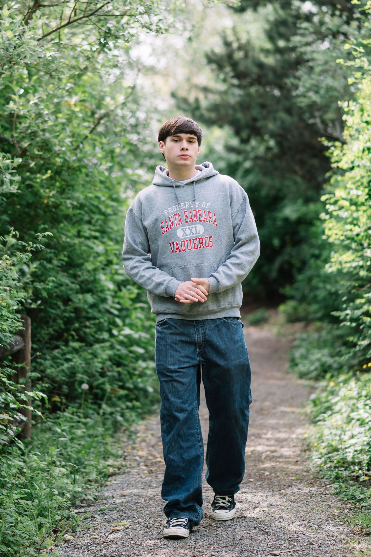 Teen boy in gray university hoodie and jeans walking on a tree-lined path outdoors.