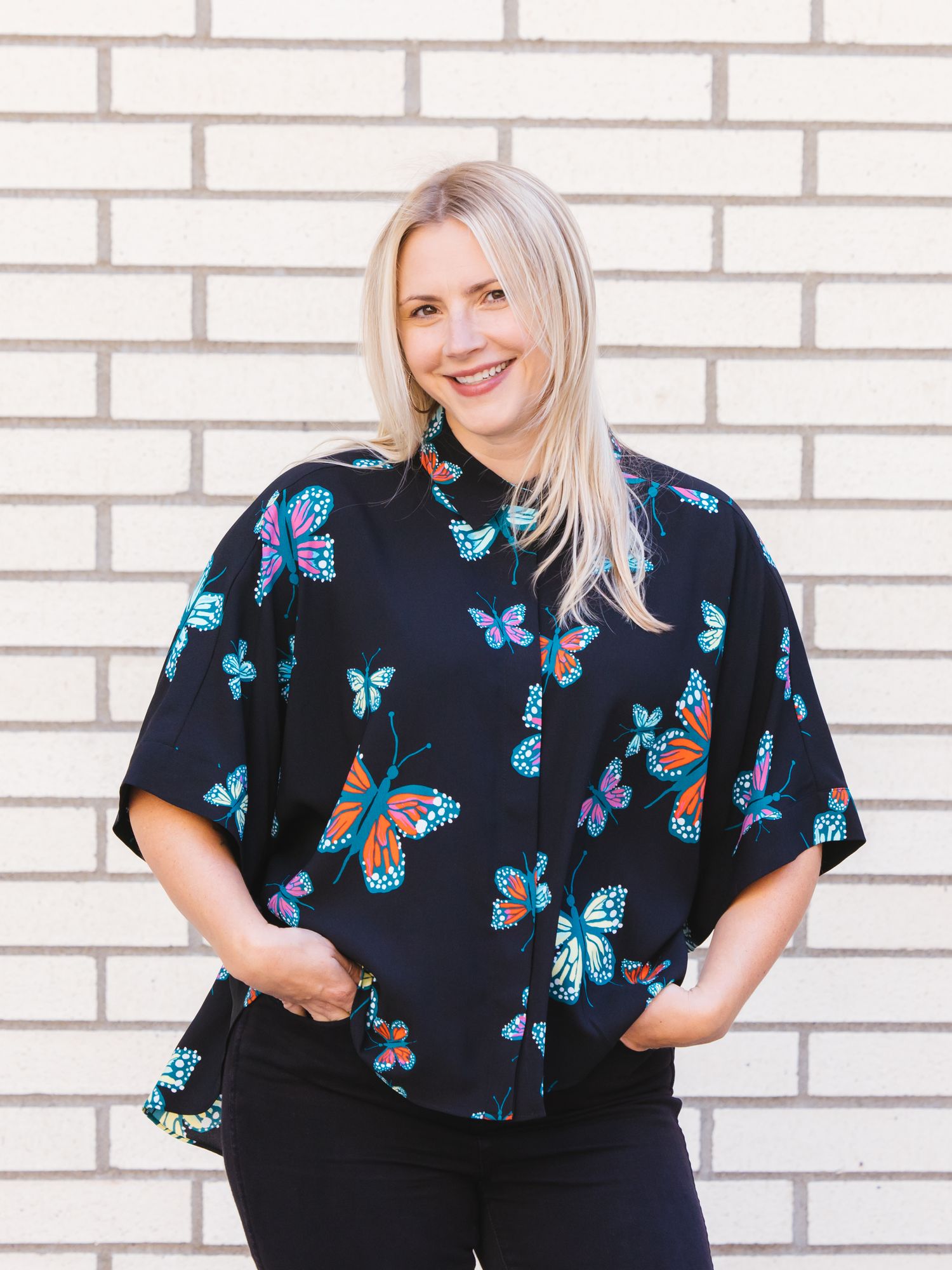 Blonde woman wearing black butterfly print blouse poses against white brick wall.