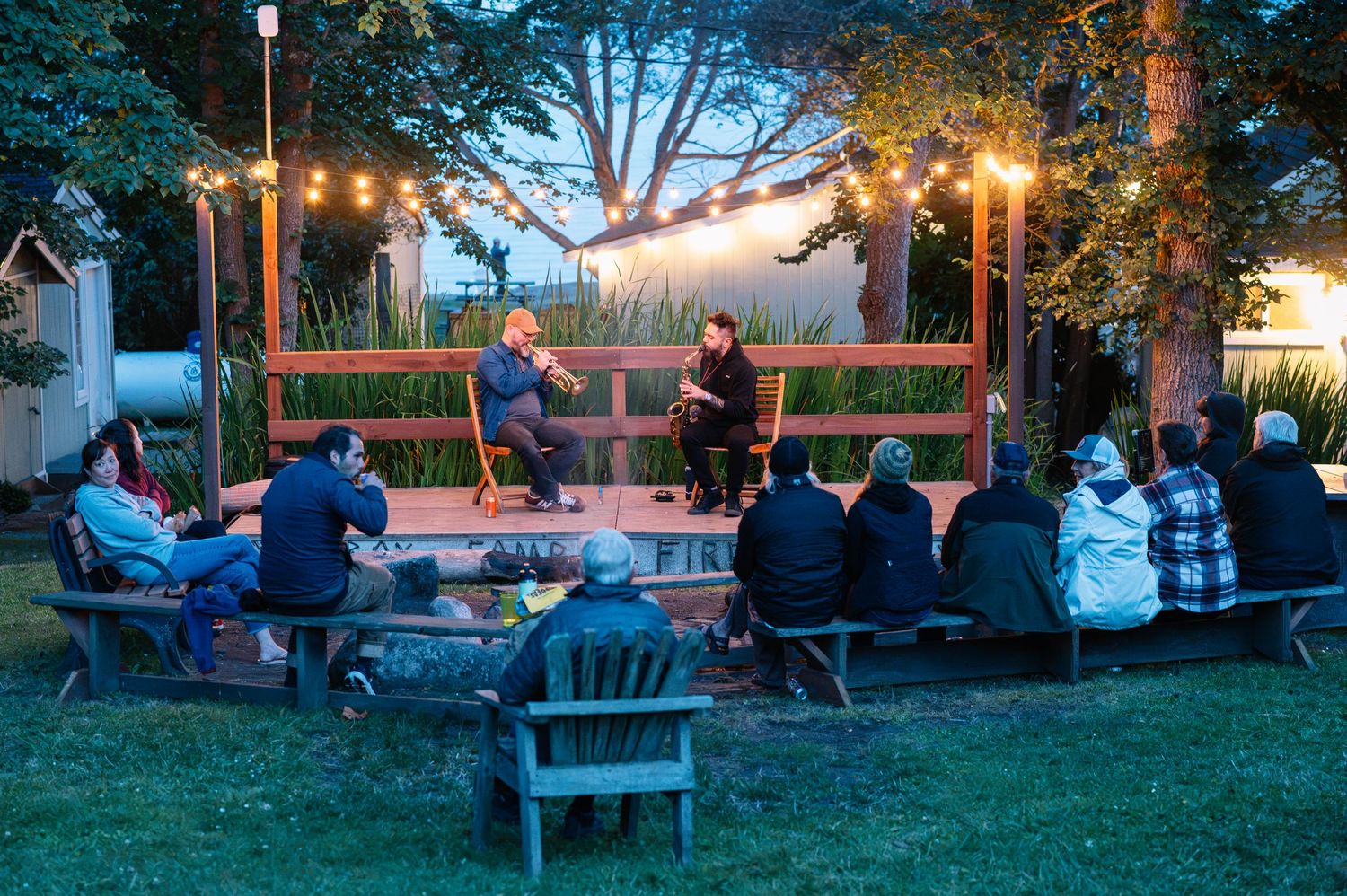 Outdoor evening gathering with audience on benches watching two speakers on a wooden stage adorned with string lights.