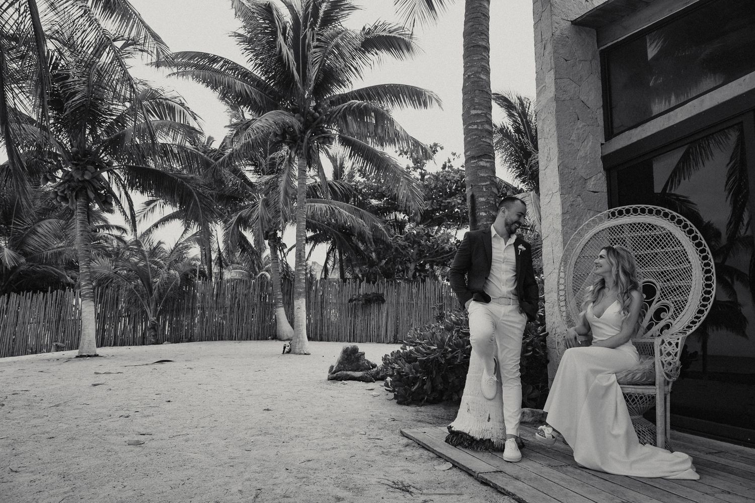 Black and white beach wedding photo of groom kissing bride seated in ornate peacock chair.