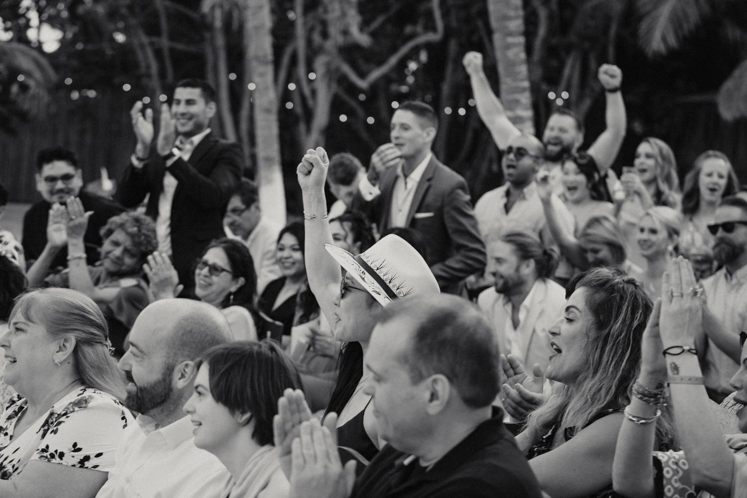 Wedding guests cheer and celebrate with raised fists during outdoor tropical wedding ceremony in black and white photo.