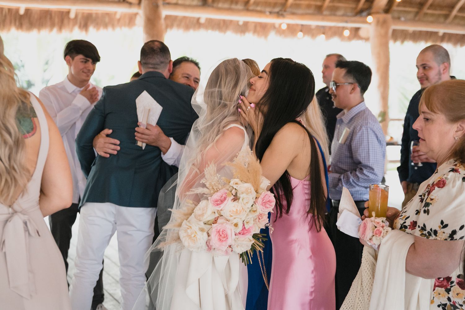 Joyful bride holding pink bouquet receives warm hugs from guests during wedding reception celebration.