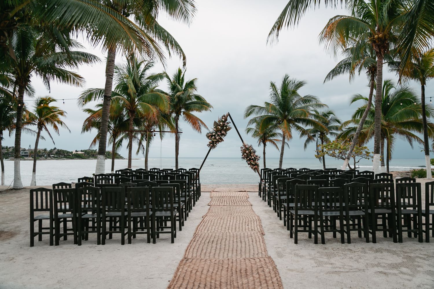 Beach wedding ceremony setup with black chairs, brick aisle, floral arch, palm trees, and ocean view.