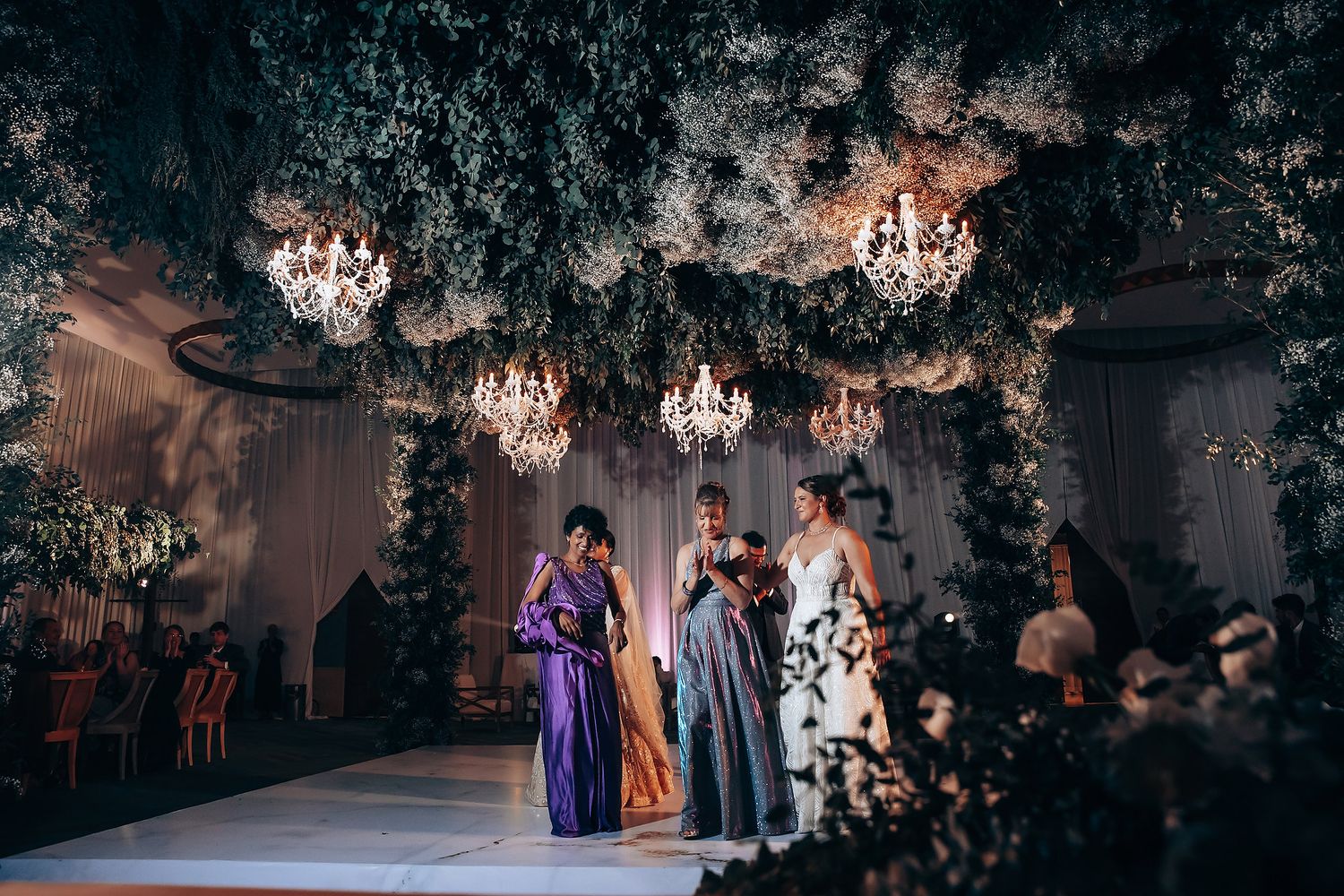 Wedding celebration under crystal chandeliers and lush greenery with guests in formal attire.