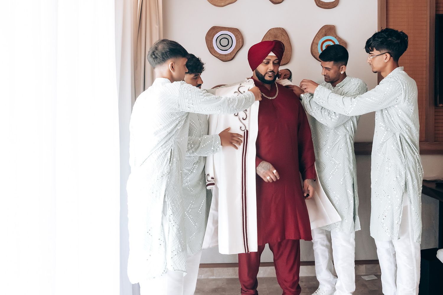 Groomsmen in white helping adjust the groom's maroon traditional outfit.
