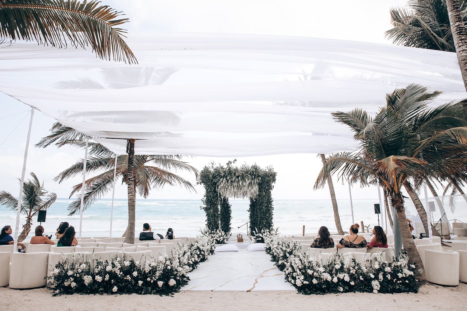 Beachfront wedding setup with white draping and floral arch against ocean backdrop.