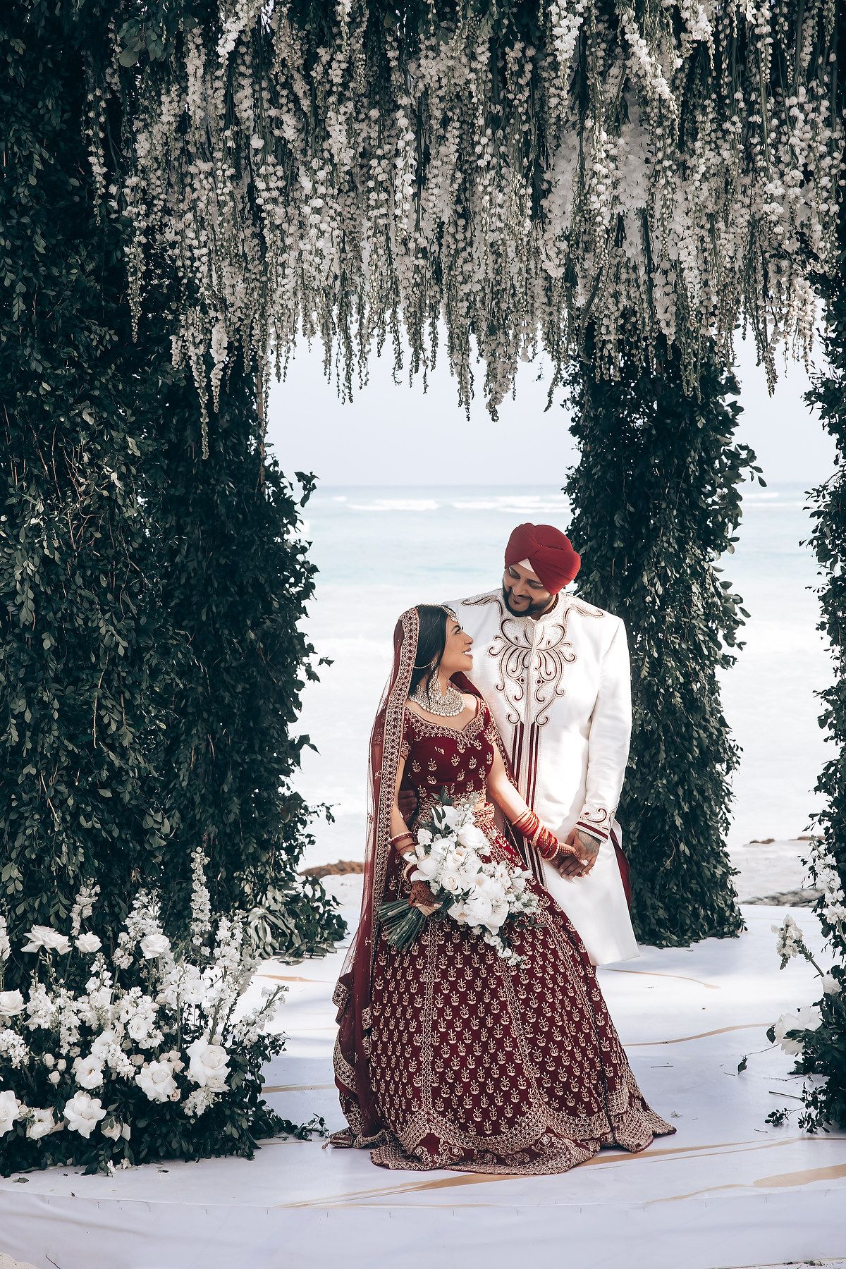 Wedding couple in burgundy and white attire pose under elegant floral arrangements.