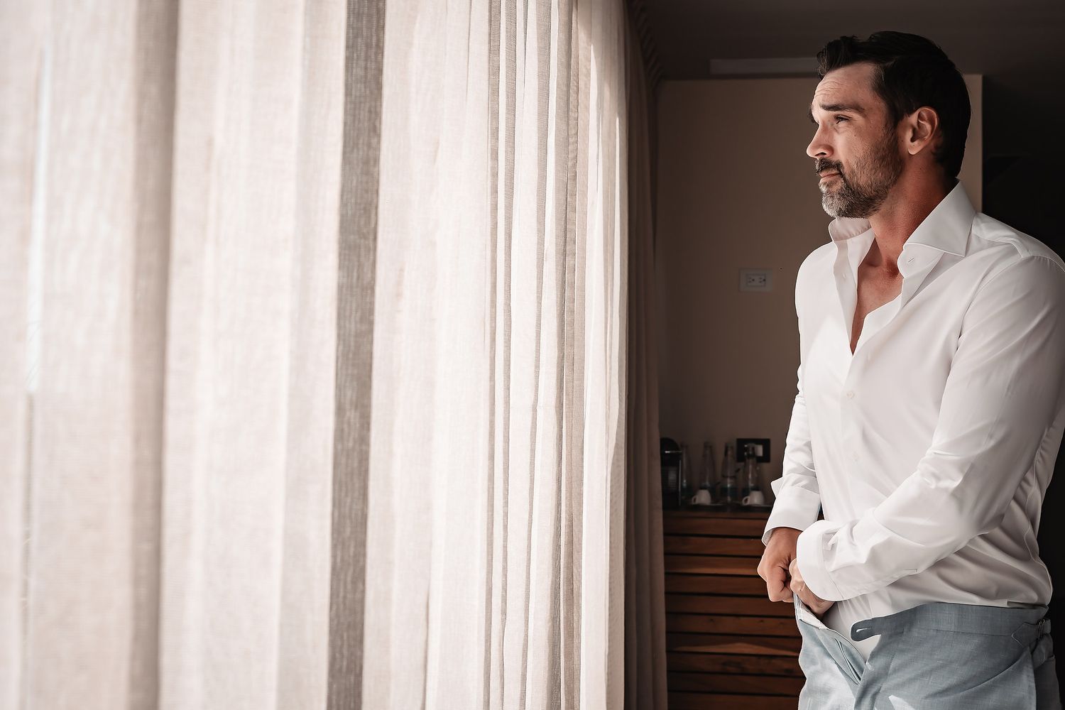Natural light portraits of groom preparation by window in classic white shirt.