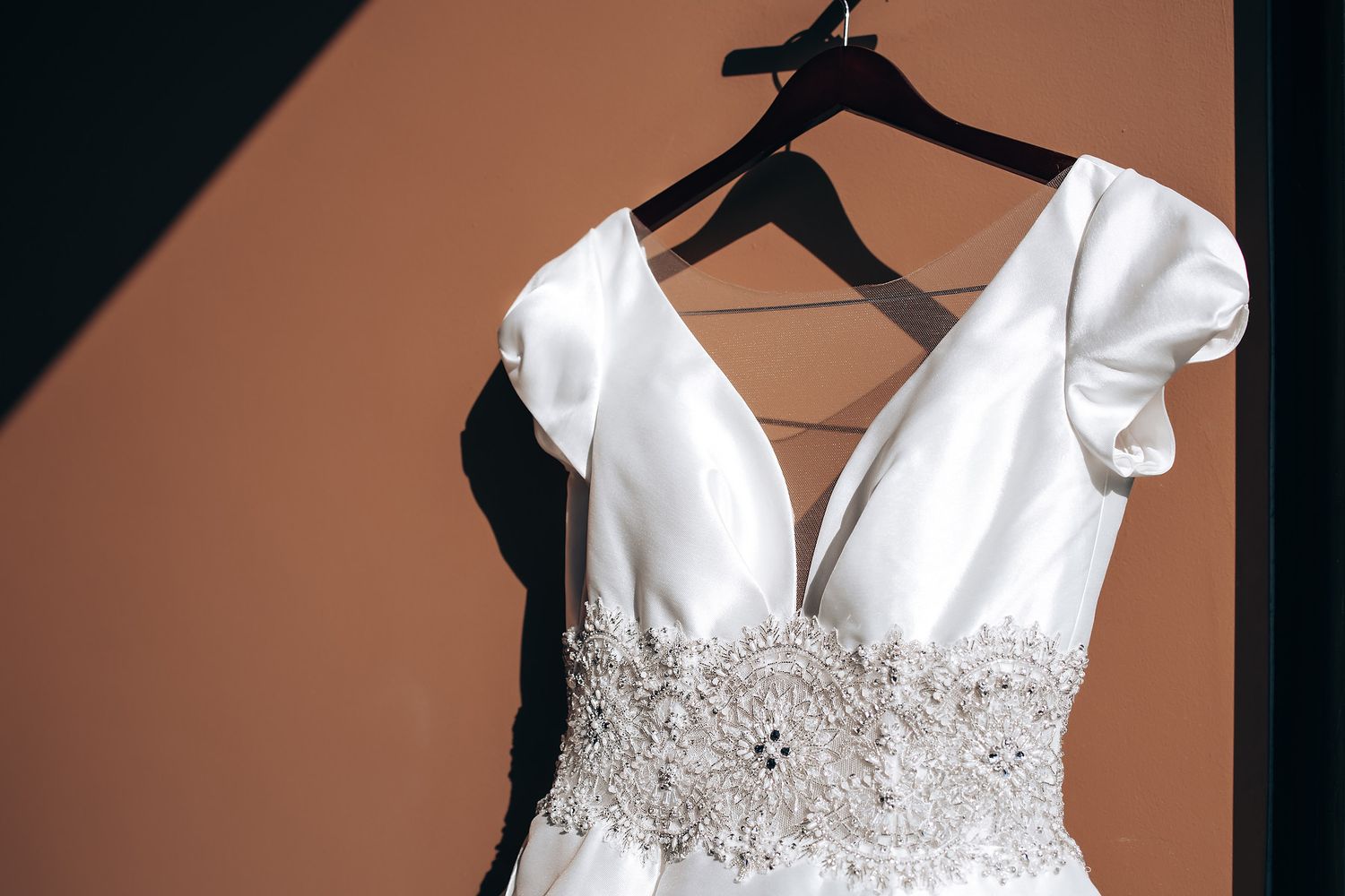 An elegant white wedding dress with beaded waistband detail hangs on display against a brown background.