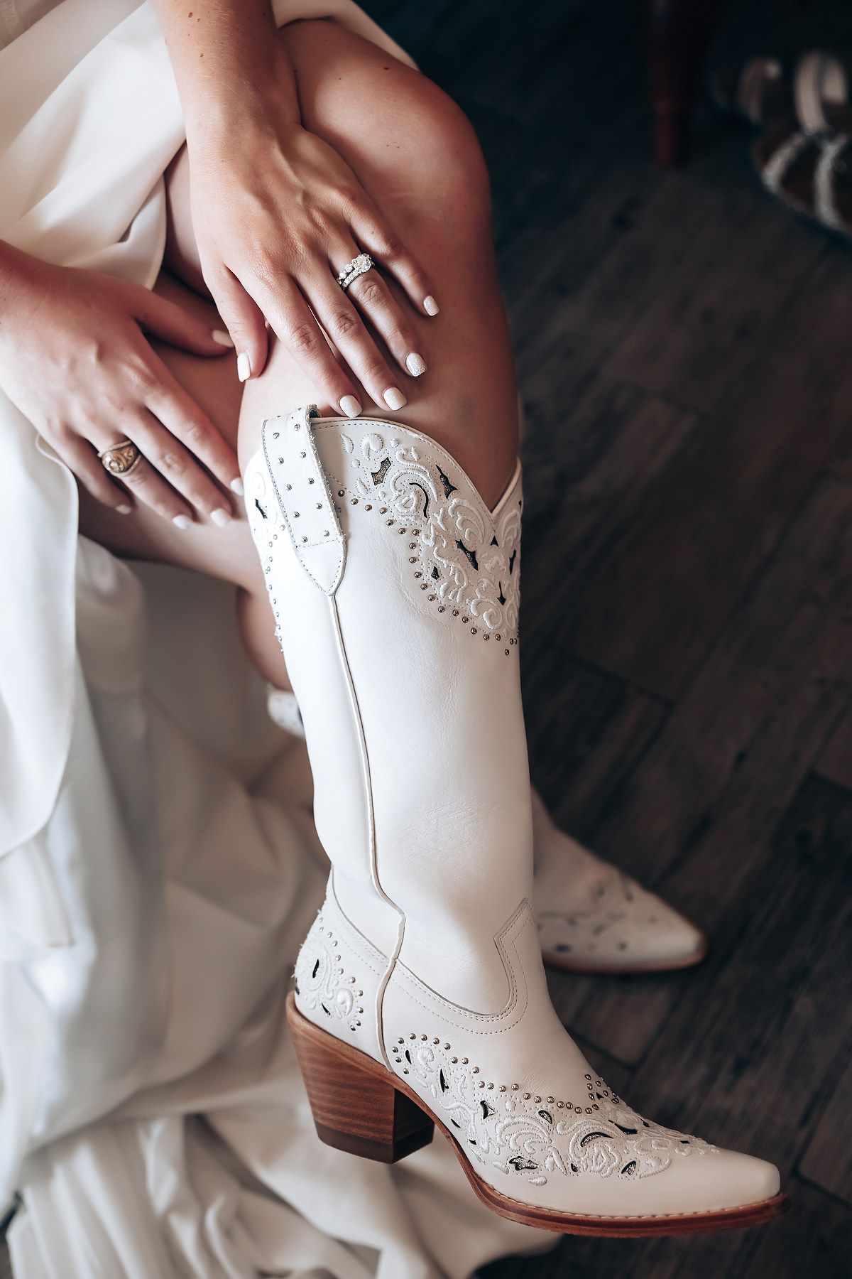 White cowboy boots with intricate cutout designs are showcased against a dark wooden floor and white dress fabric.