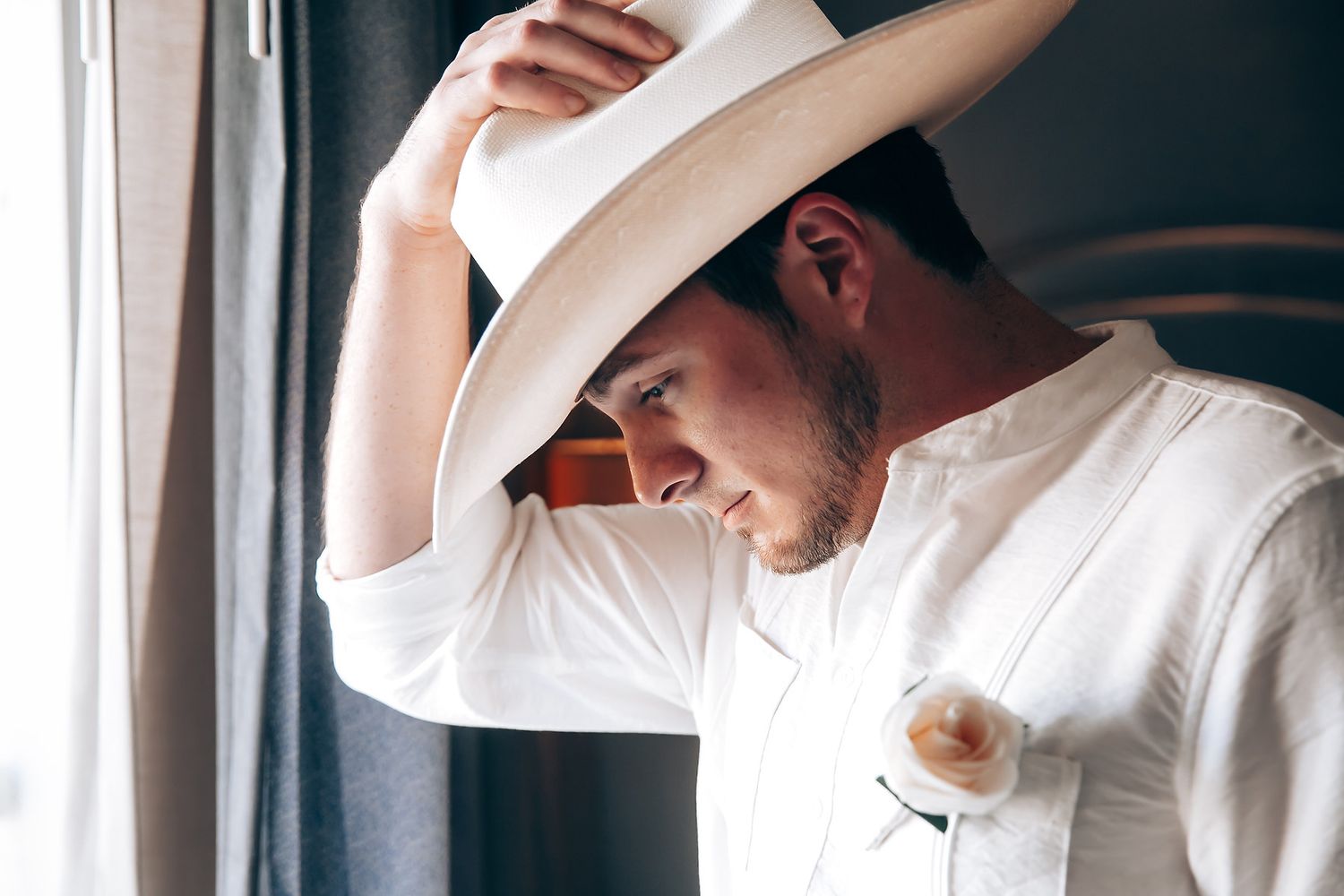 A person wearing a white cowboy hat and white button-up shirt leans against a wall in a dimly lit room.