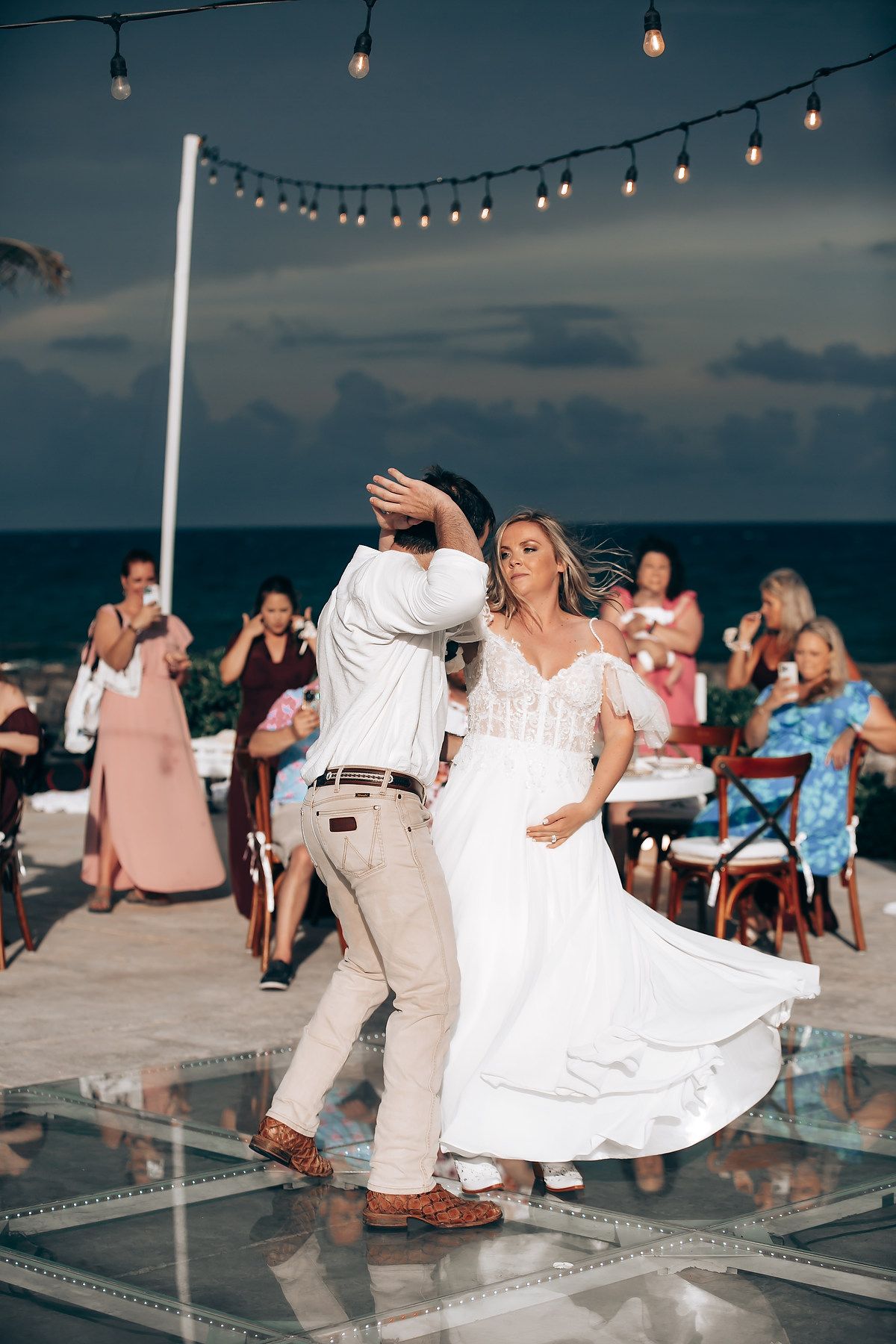 Couple dancing on a beachside deck at sunset with string lights overhead.