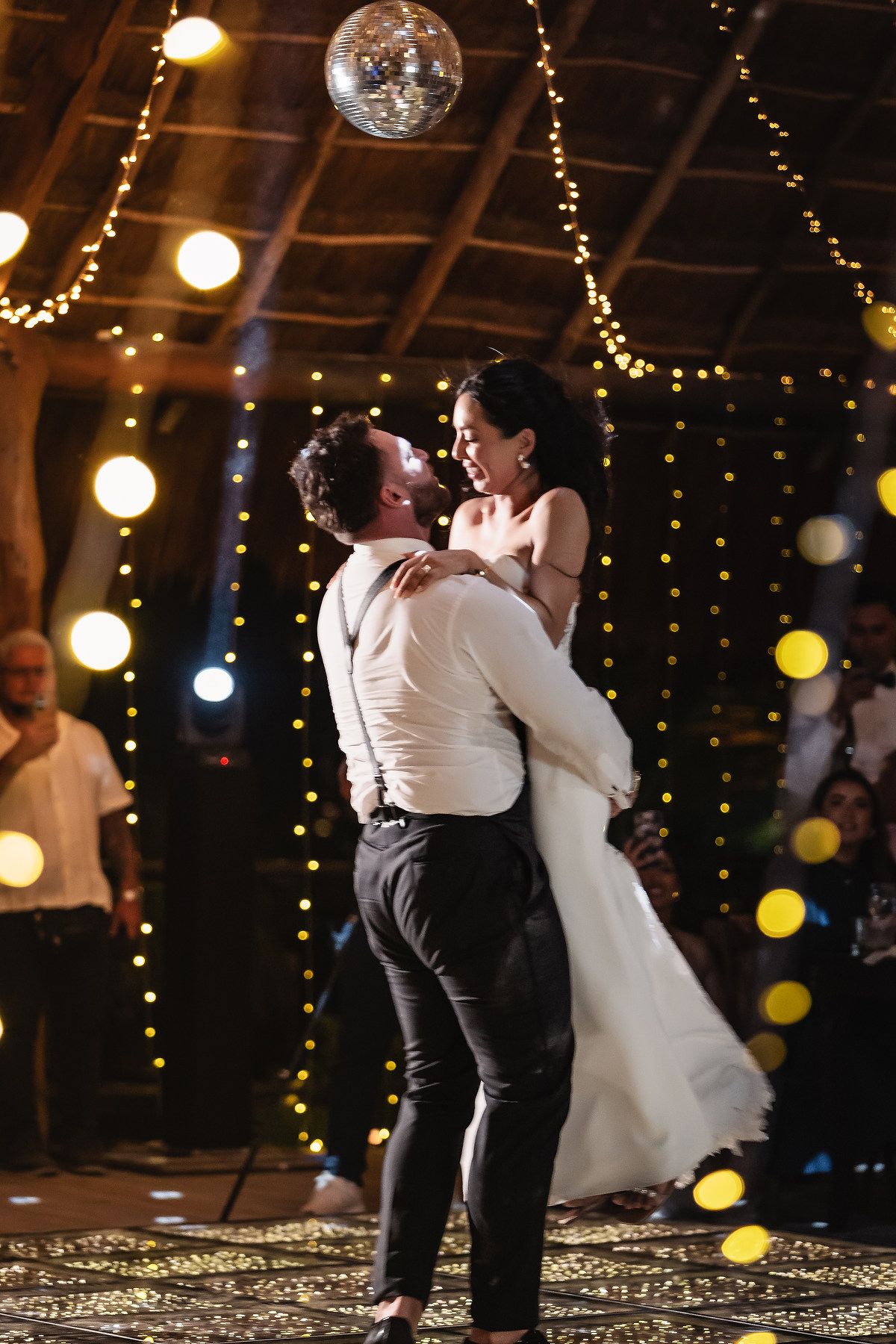 A couple dances together on a patterned floor under warm barn lighting and disco balls.