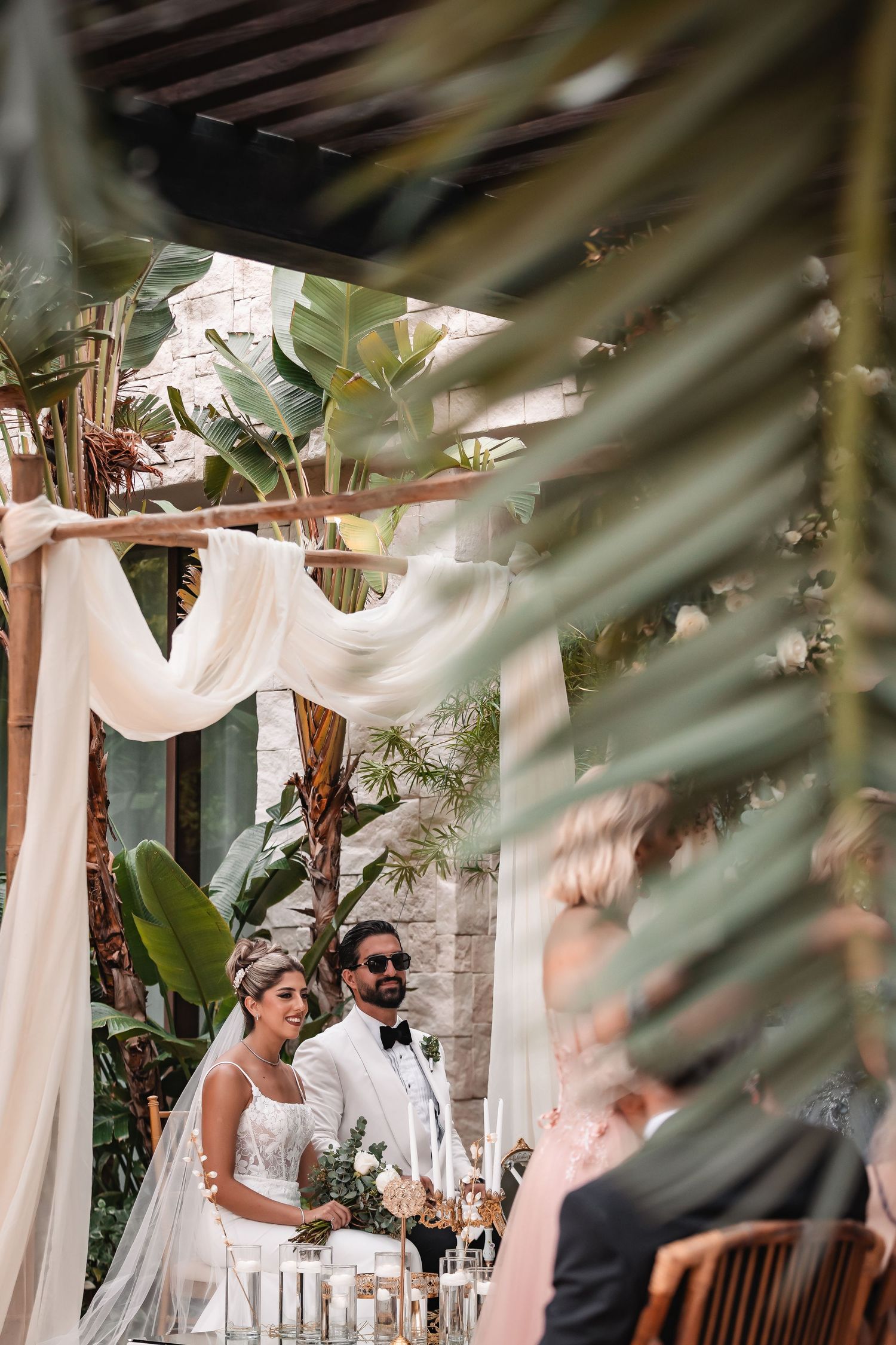 Elegant outdoor wedding ceremony through tropical foliage with white fabric draping.
