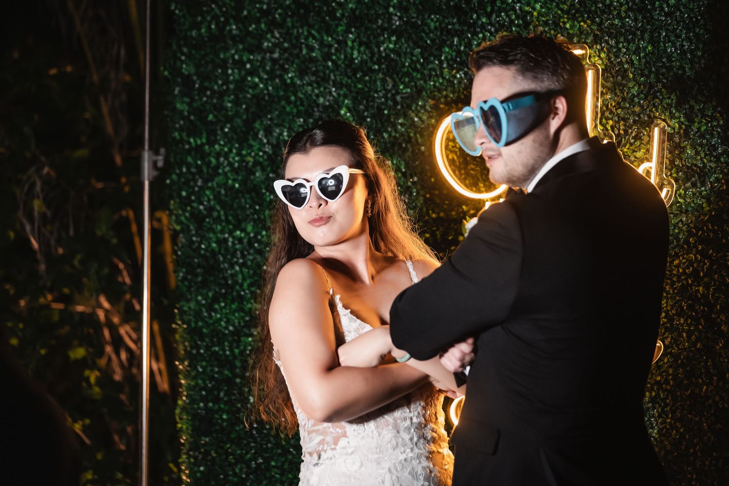 A stylish couple wearing sunglasses poses for nighttime wedding photos with illuminated greenery.