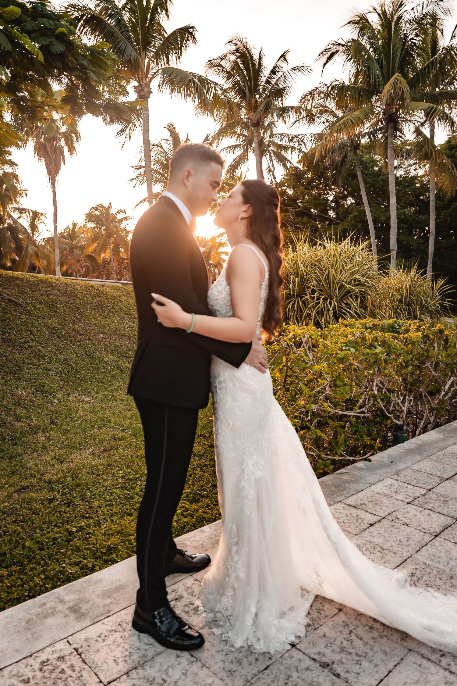 Newlyweds share intimate moment at sunset with tropical palm trees in background.