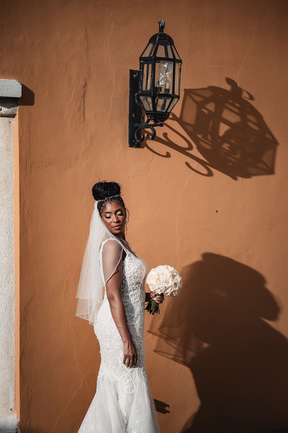 A bride in a white lace dress poses against a peach wall with dramatic lantern shadows.