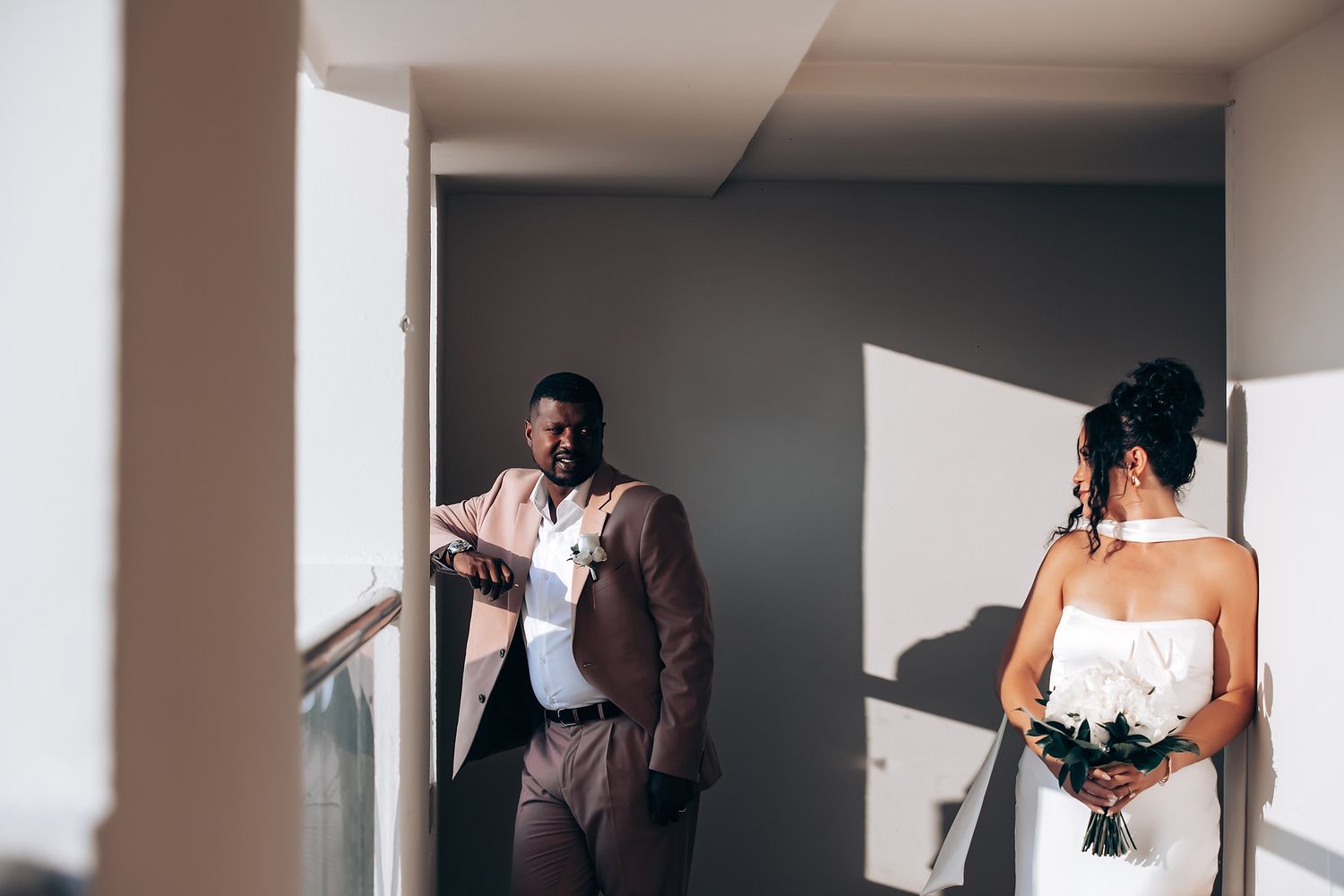 Wedding party members in formal attire stand in sunlit hallway with dramatic shadows.