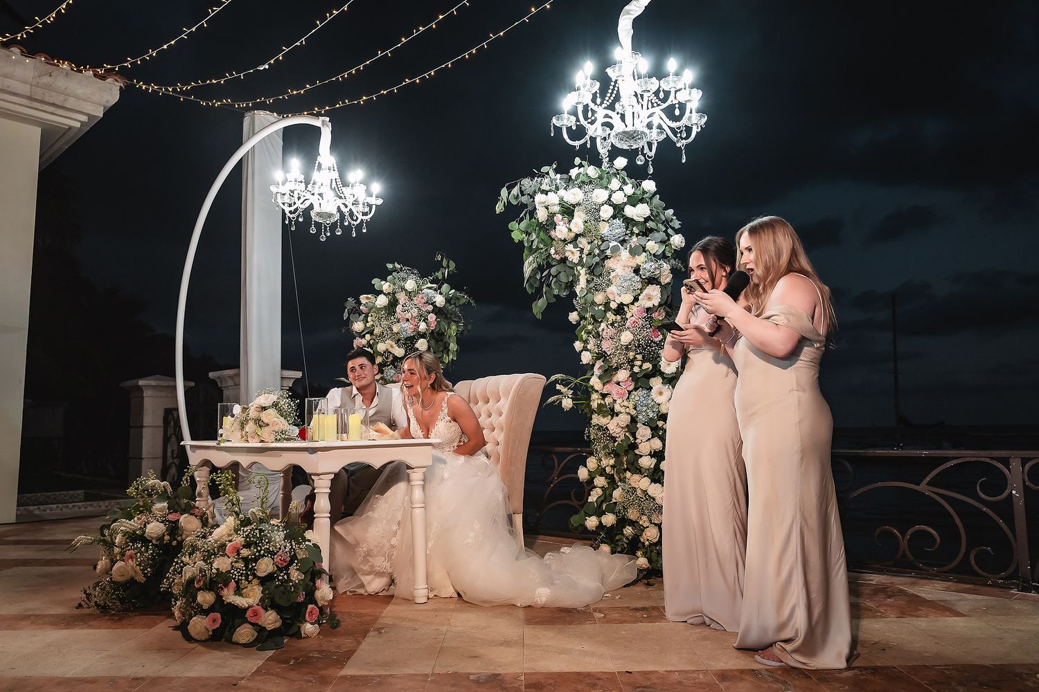 Elegant wedding reception setup with crystal chandeliers and floral arrangements on a terrace at night.